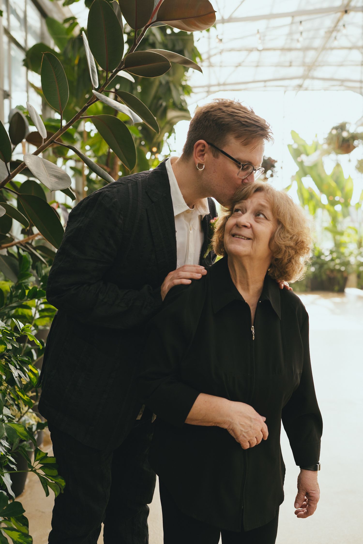 Young man in dark suit kissing older smiling woman on cheek among lush green plants in greenhouse setting.