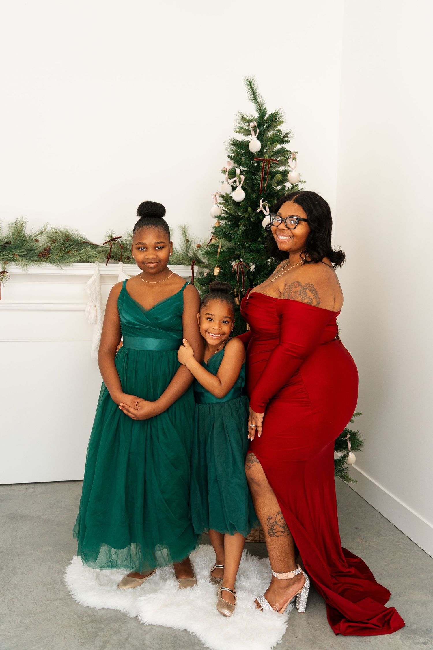 A mother in red gown poses with two daughters in green dresses by a Christmas tree.