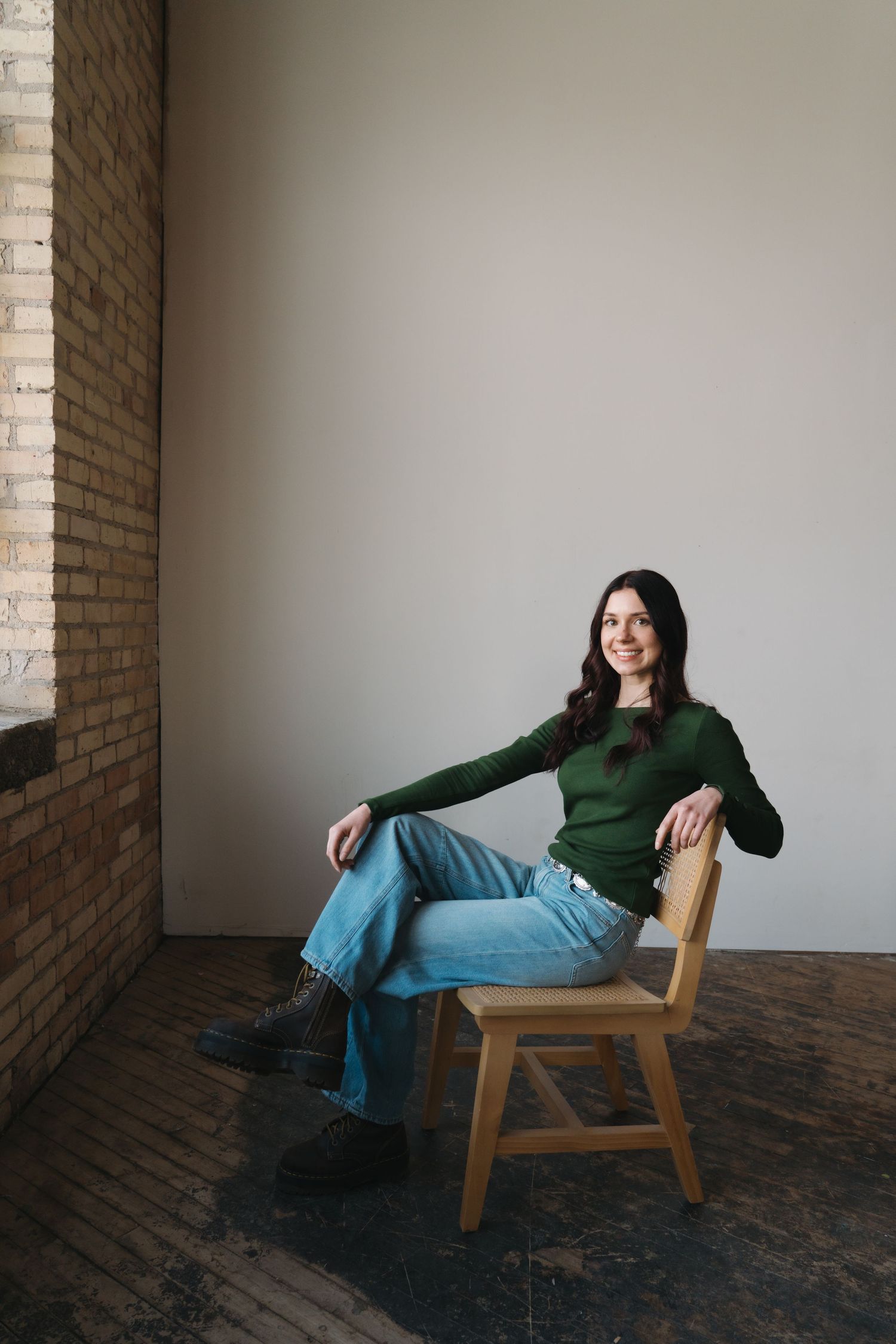 A woman in a green sweater and jeans sits relaxed on a wooden chair near a brick wall.