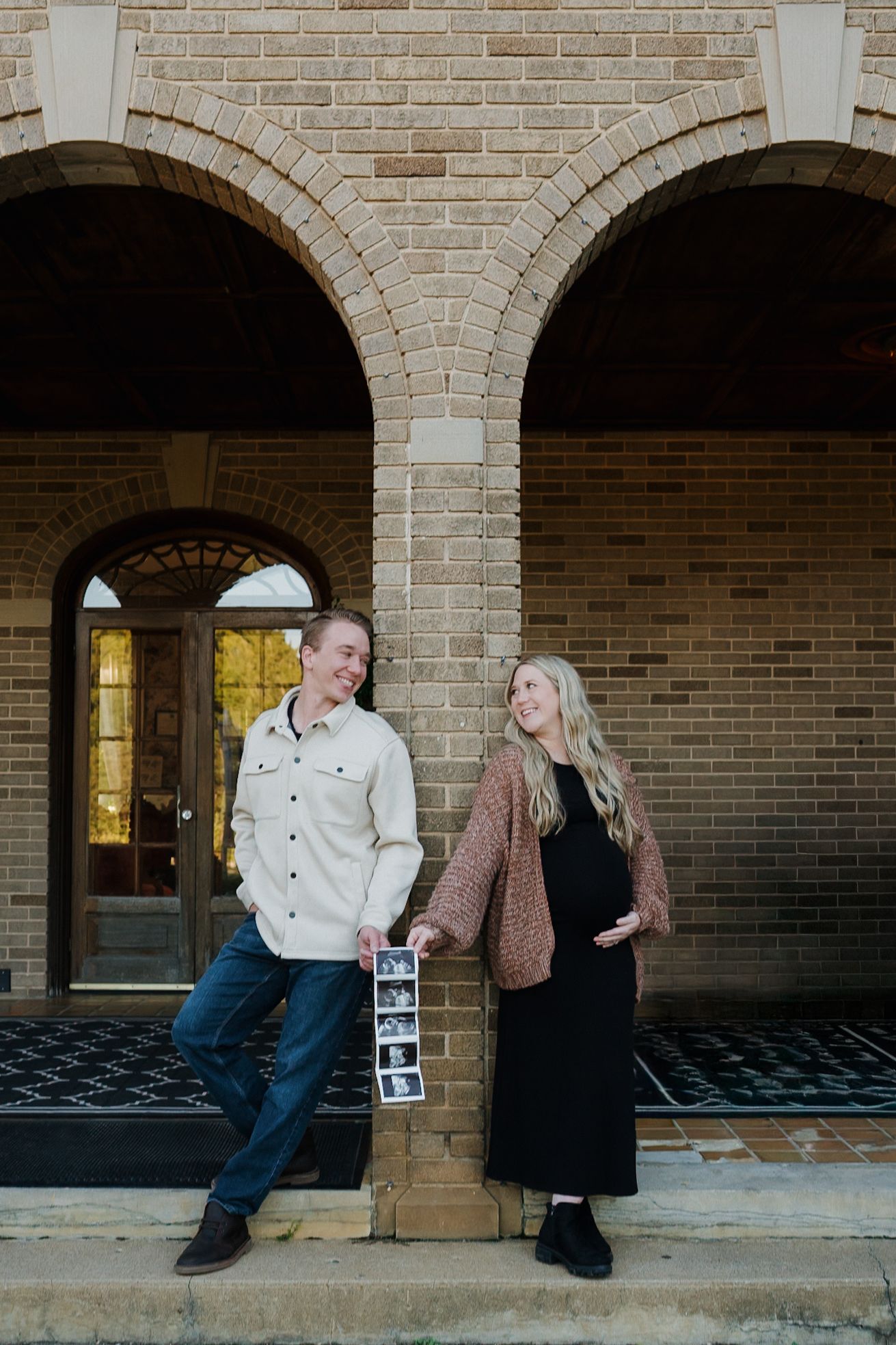 A couple holds ultrasound photos while posing before a brick building during maternity shoot.