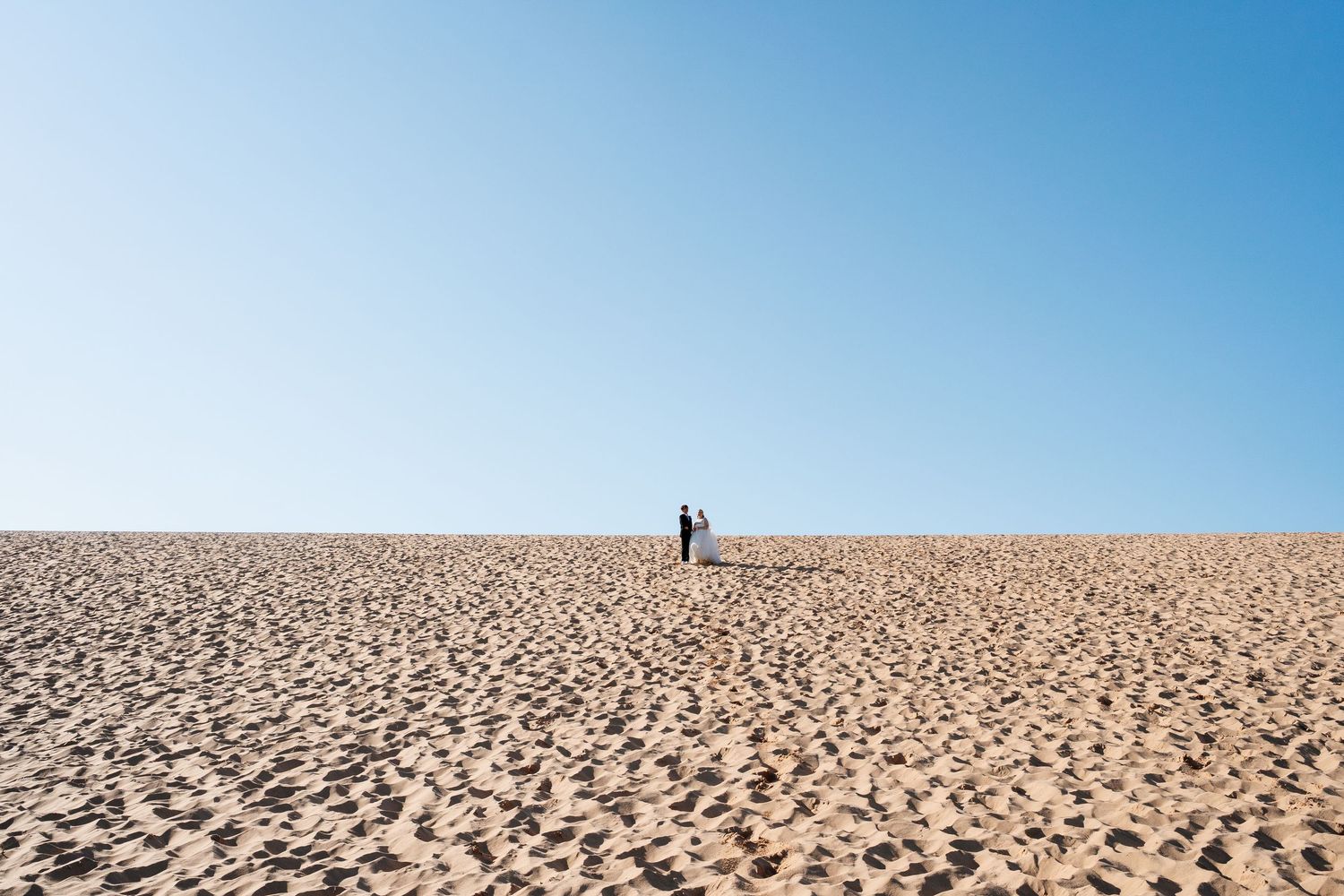 Bride and groom standing together atop a vast sandy dune under clear blue sky.