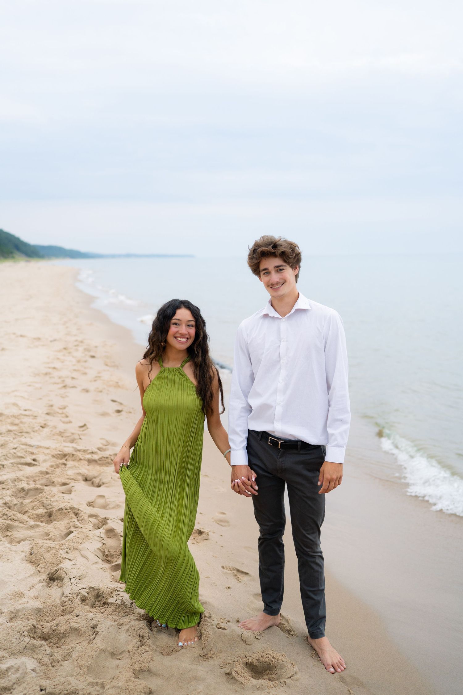 A young couple holds hands smiling on a sandy beach, woman in green dress, man in white shirt and black pants.