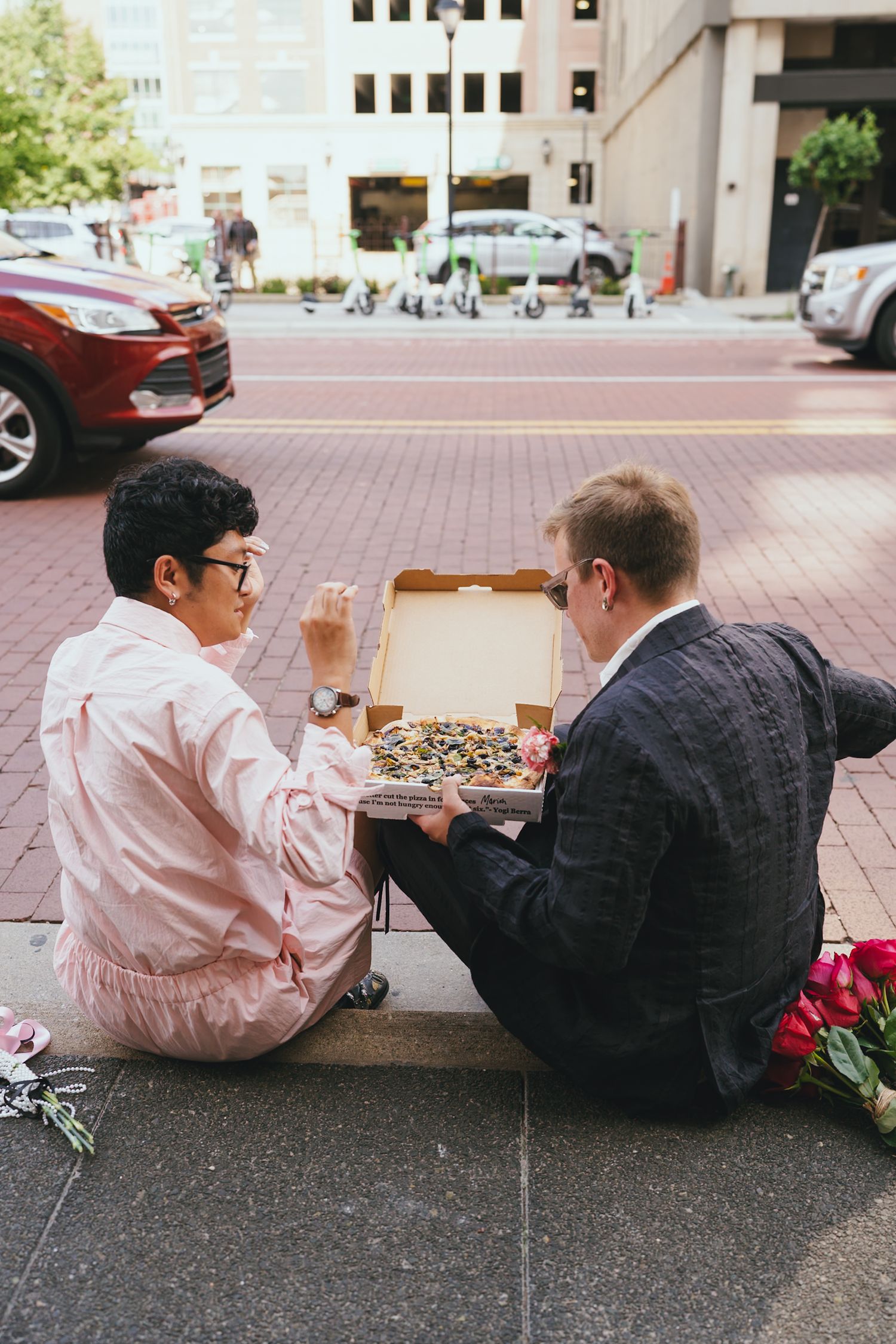 Newlywed couple sitting on city sidewalk sharing pizza after courthouse wedding ceremony.