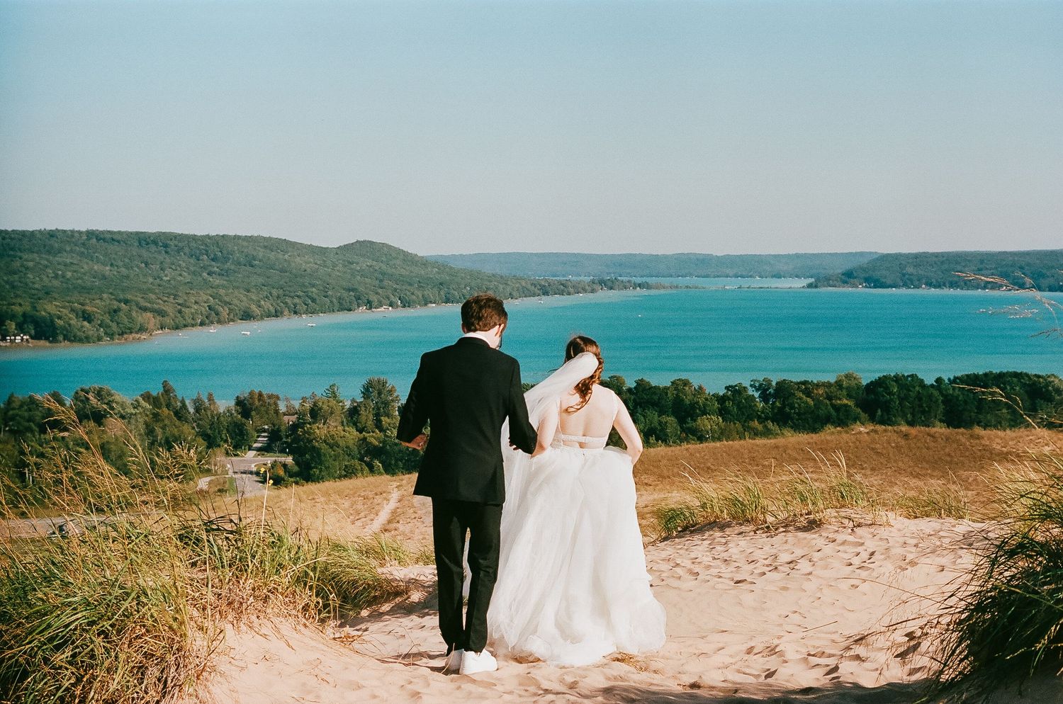 Bride and groom walking hand in hand on sandy dune overlooking turquoise lake.