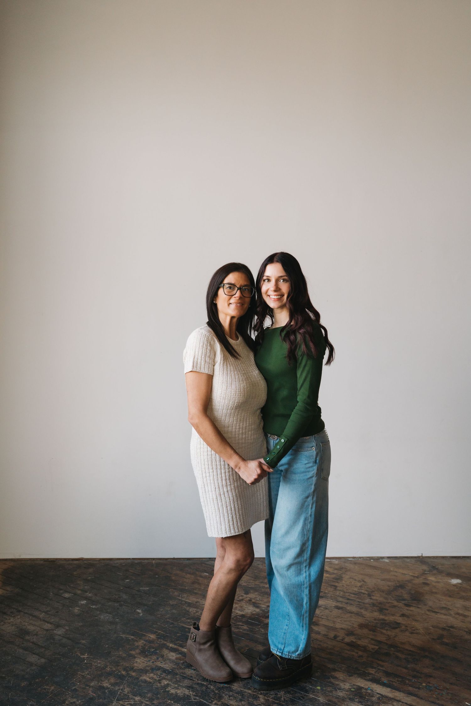 Two women smiling together in a studio, one in a beige dress with glasses, one in a green top.