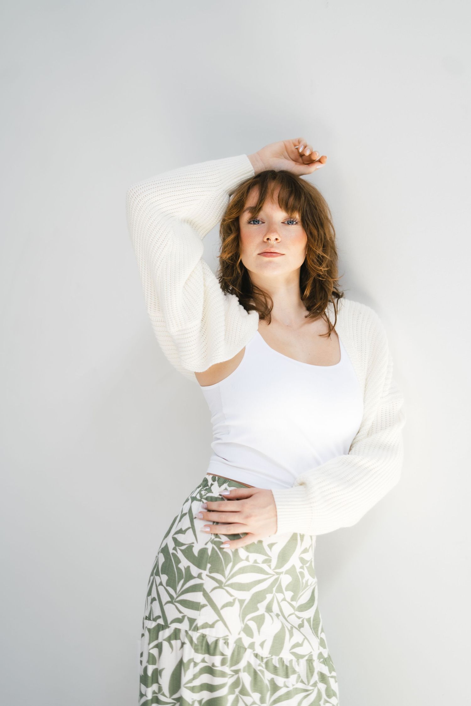 Woman in white tank top and cream knit cardigan with sage floral skirt posing with arm raised.