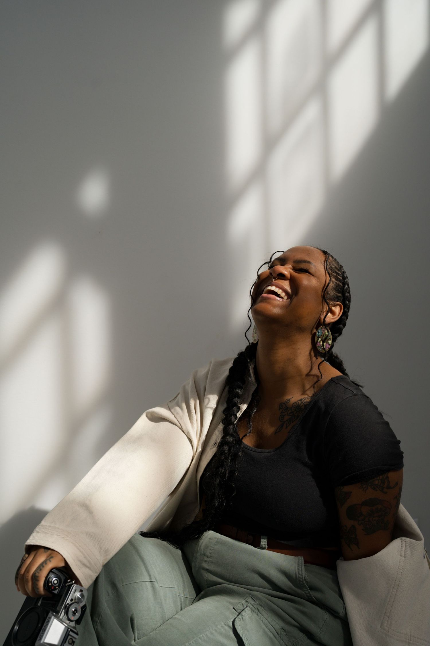 A laughing Black woman with braids sits against a grey wall in sunlight, wearing a black crop top and white jacket.