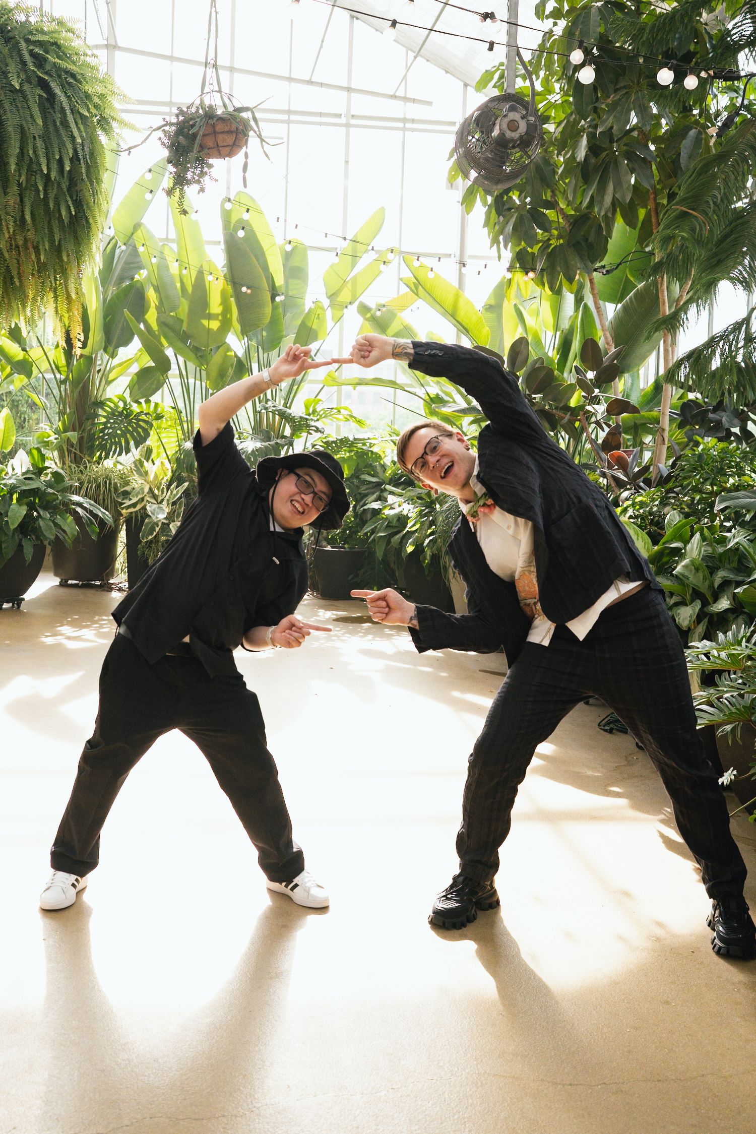Two men in black suits strike fun poses in a sunlit tropical greenhouse setting.