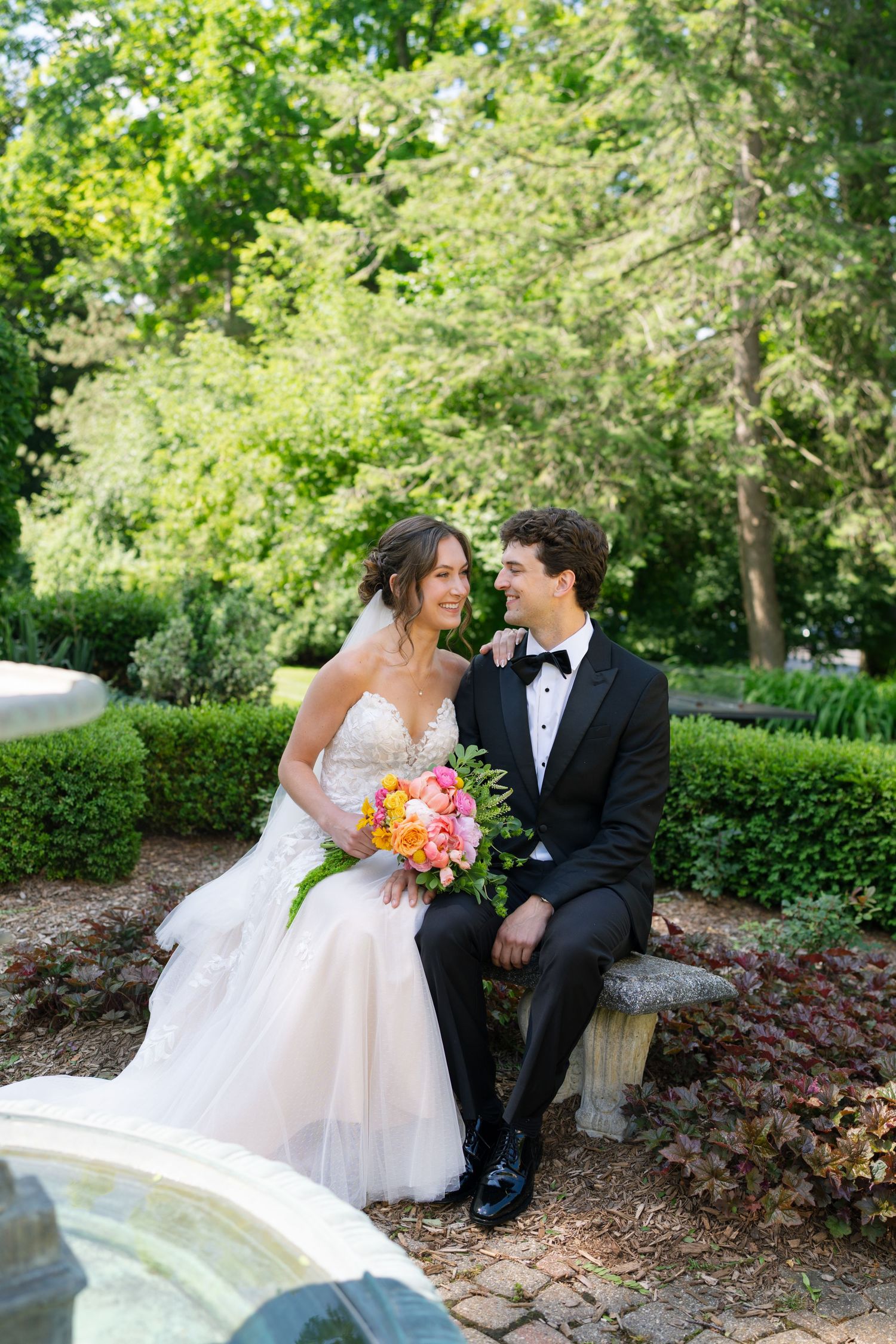 Bride and groom sit on a stone bench gazing at each other in a beautifully landscaped garden.