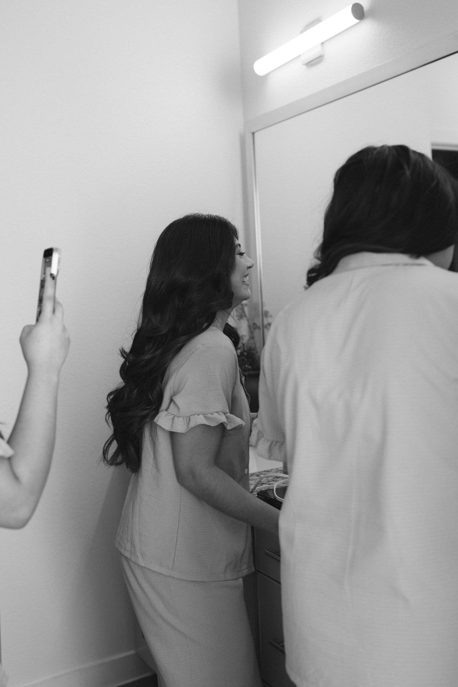 Black and white photo of bride getting ready, reflected in mirror with stylist assisting her.