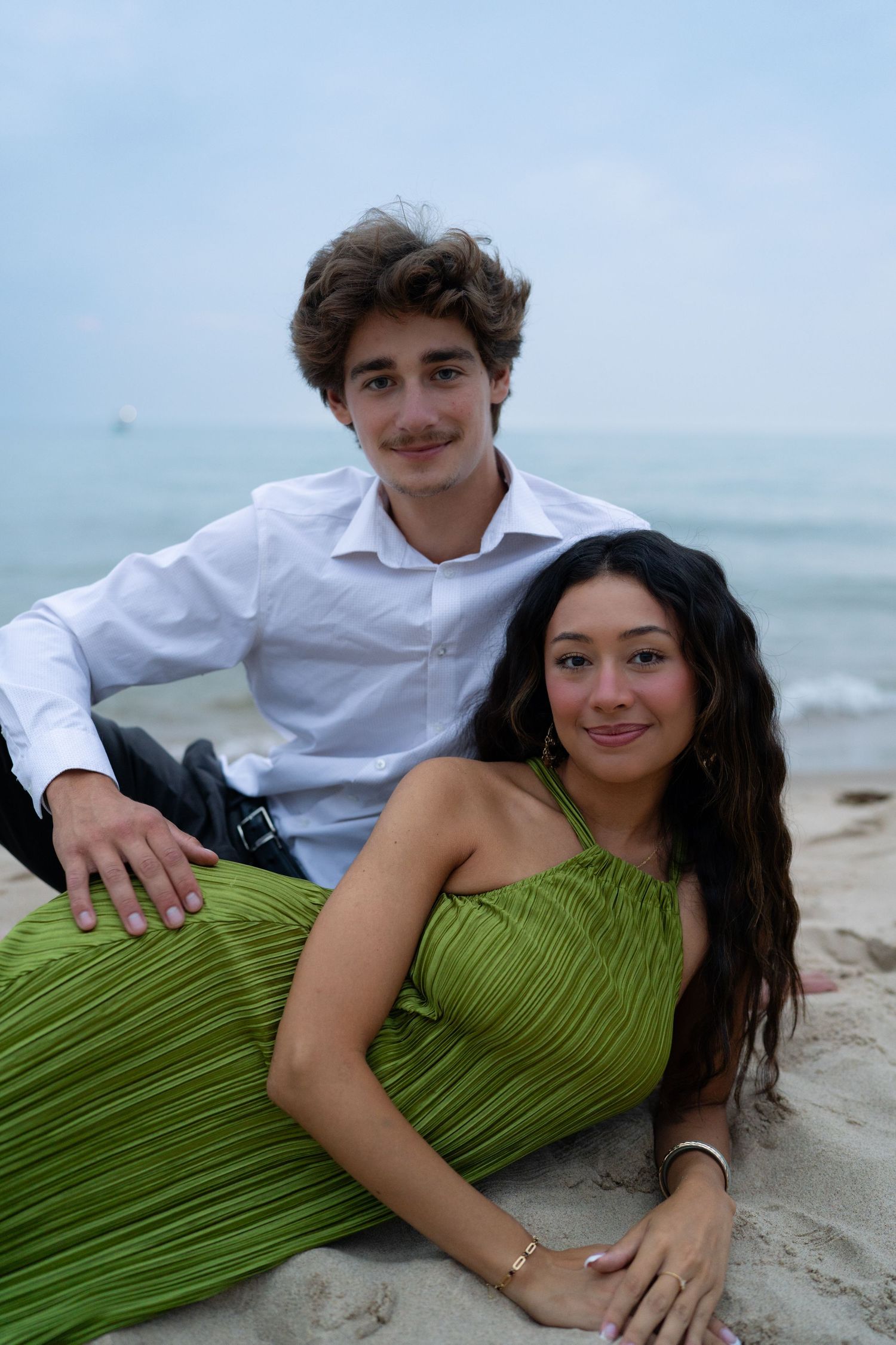 A young couple poses on a sandy beach, the woman in a green halter dress and the man in a white dress shirt.