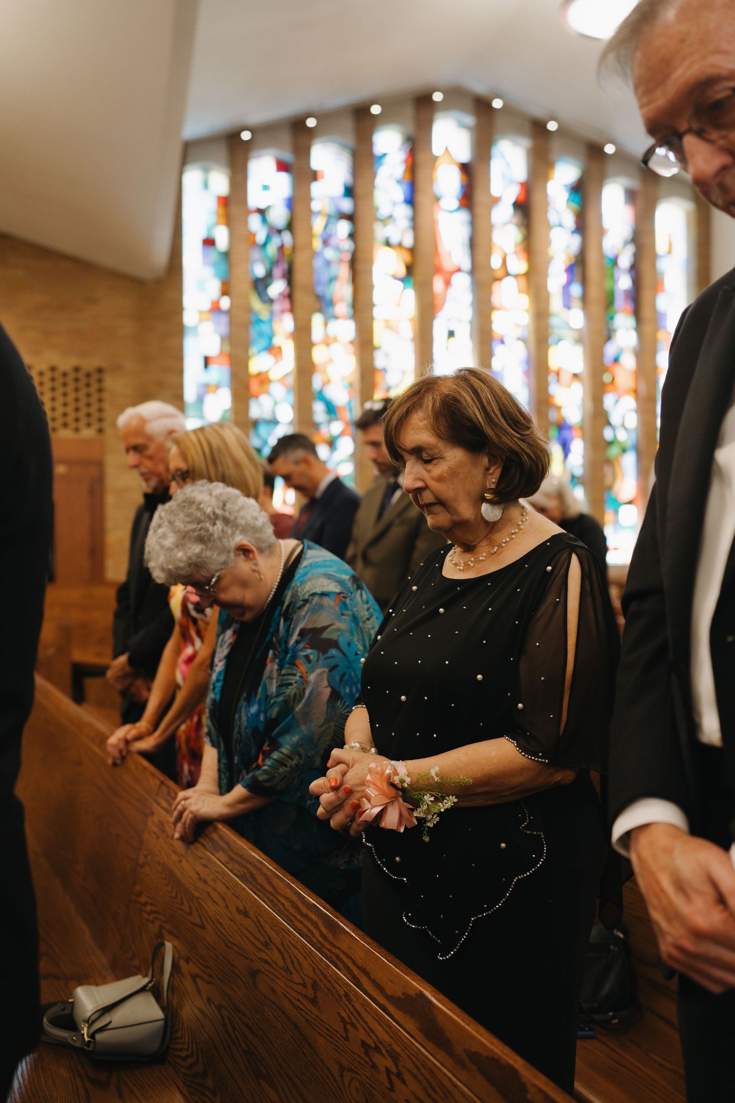 Wedding guests bow in prayer at church pews before colorful stained glass windows during ceremony.