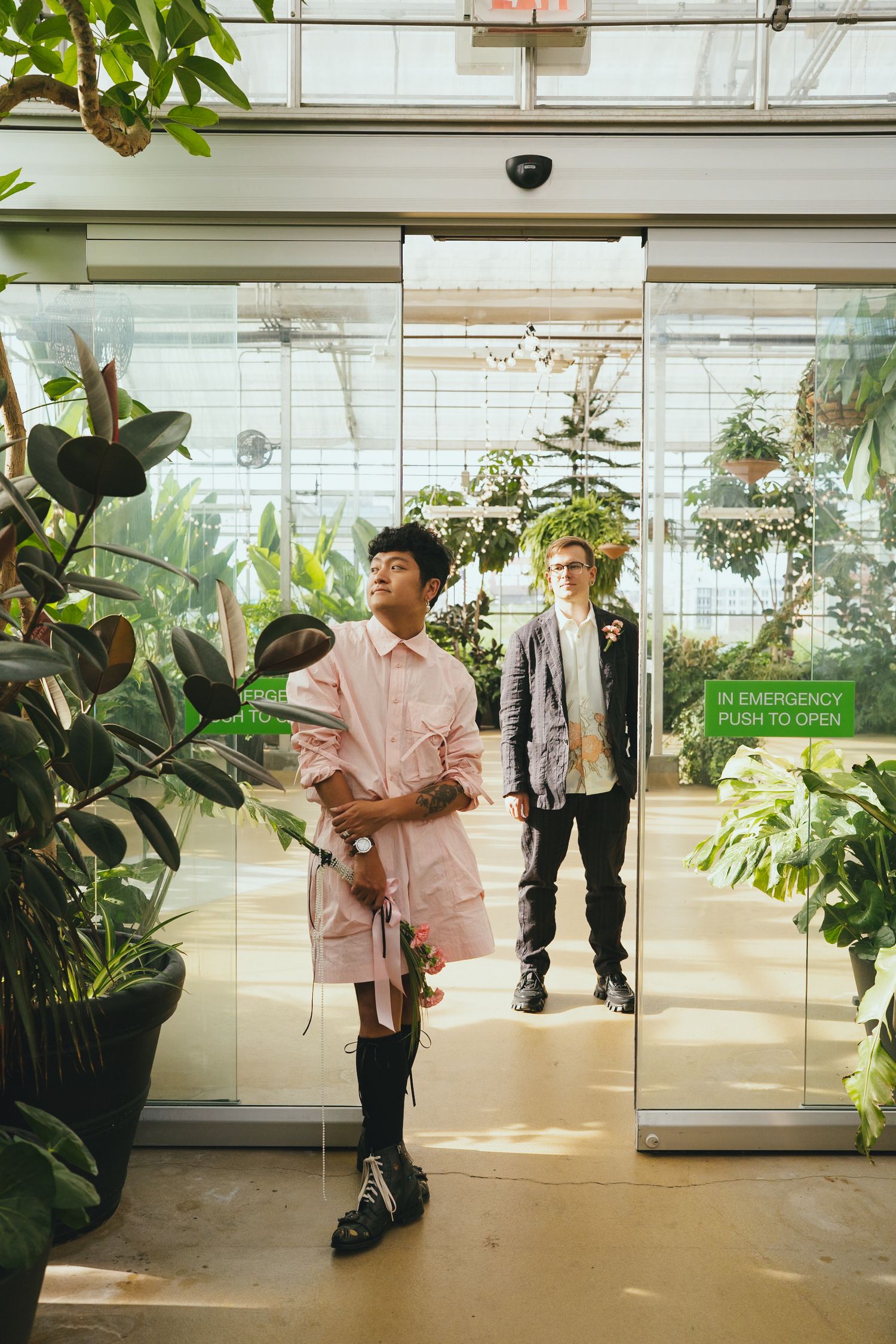 A person in a pink dress holds flowers while standing in a greenhouse glass doorway.