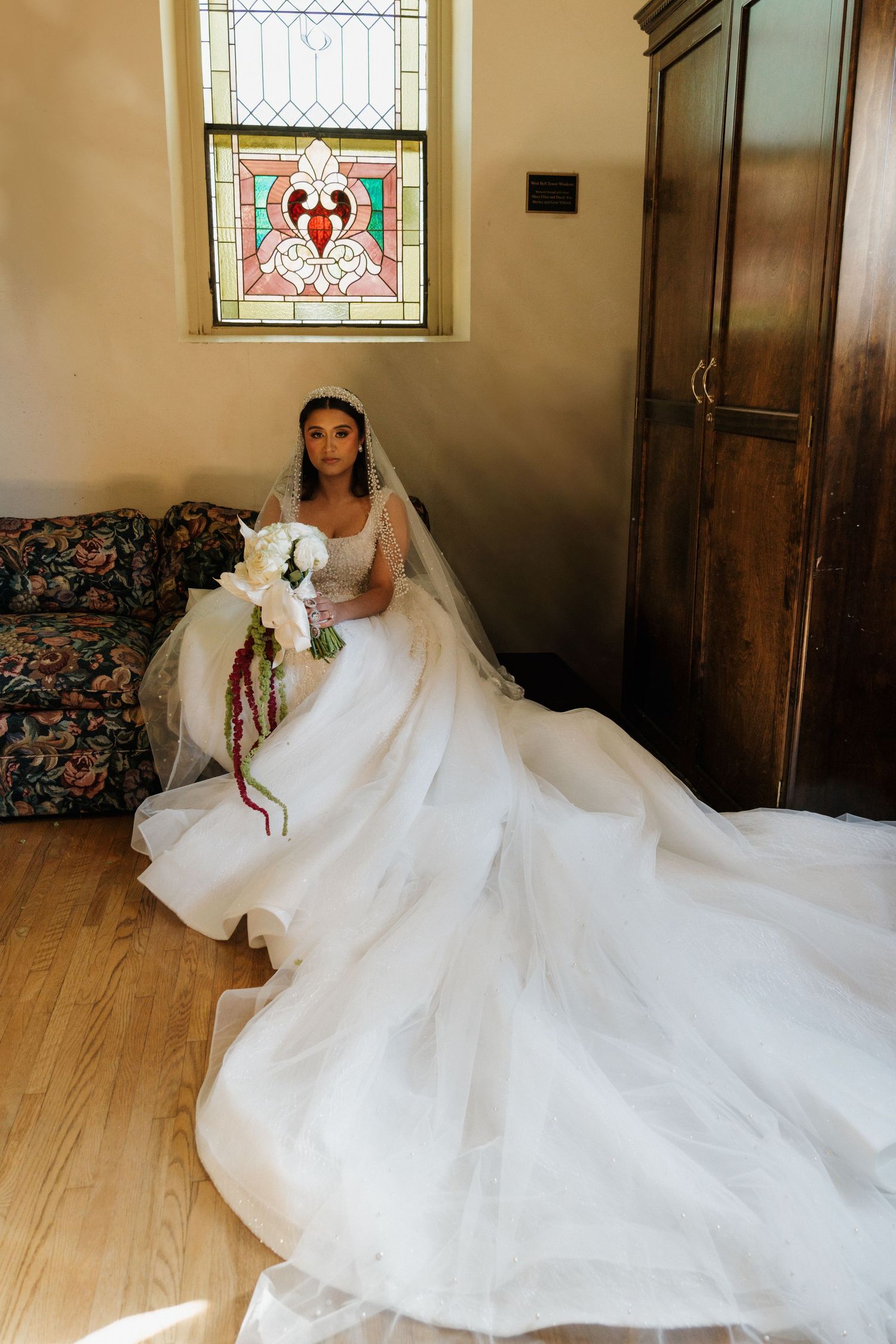 Bride in a voluminous white gown sits near a stained glass window holding a cascading bouquet.