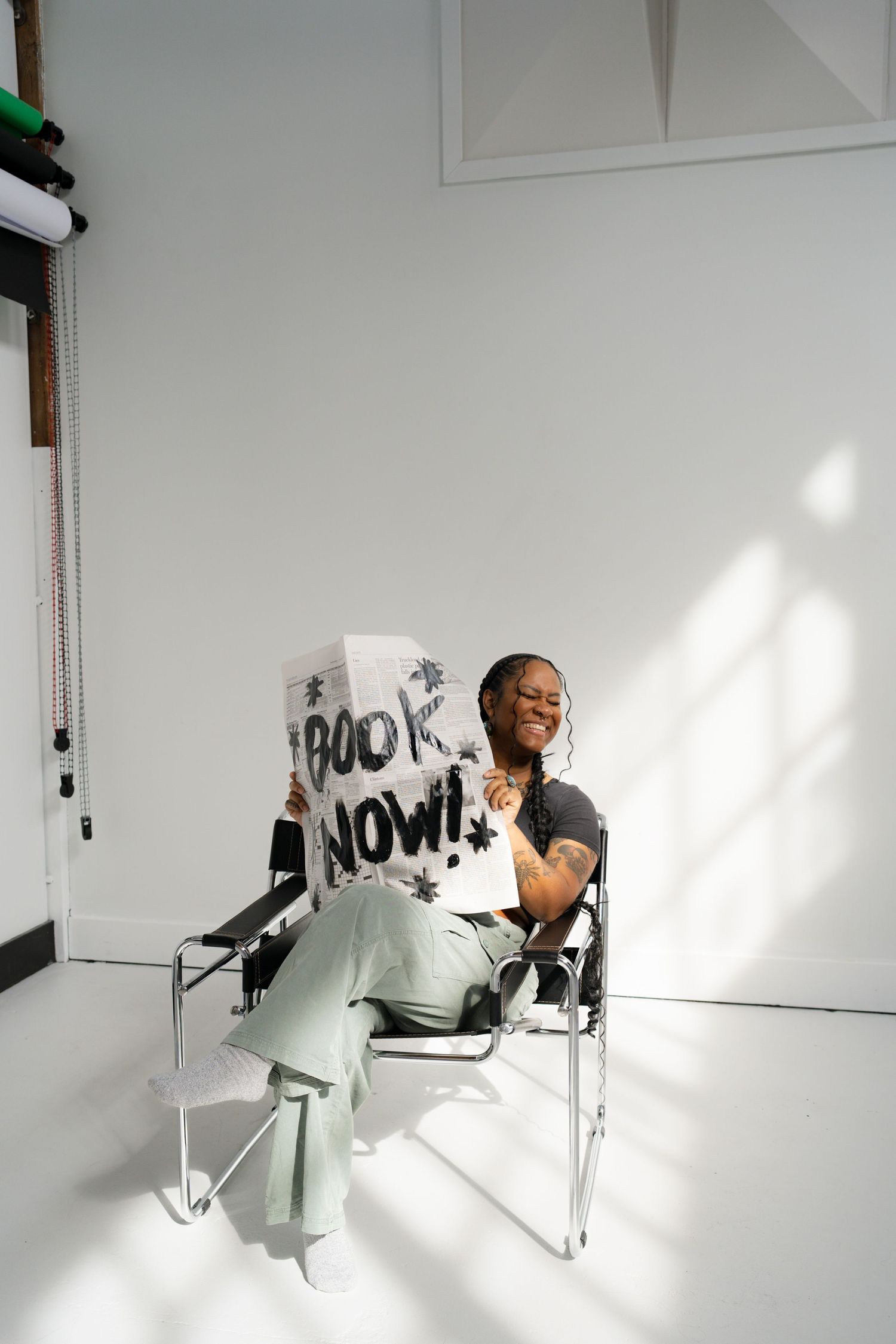Laughing person sitting in chair holding a hand-painted sign reading Book Now in white studio.