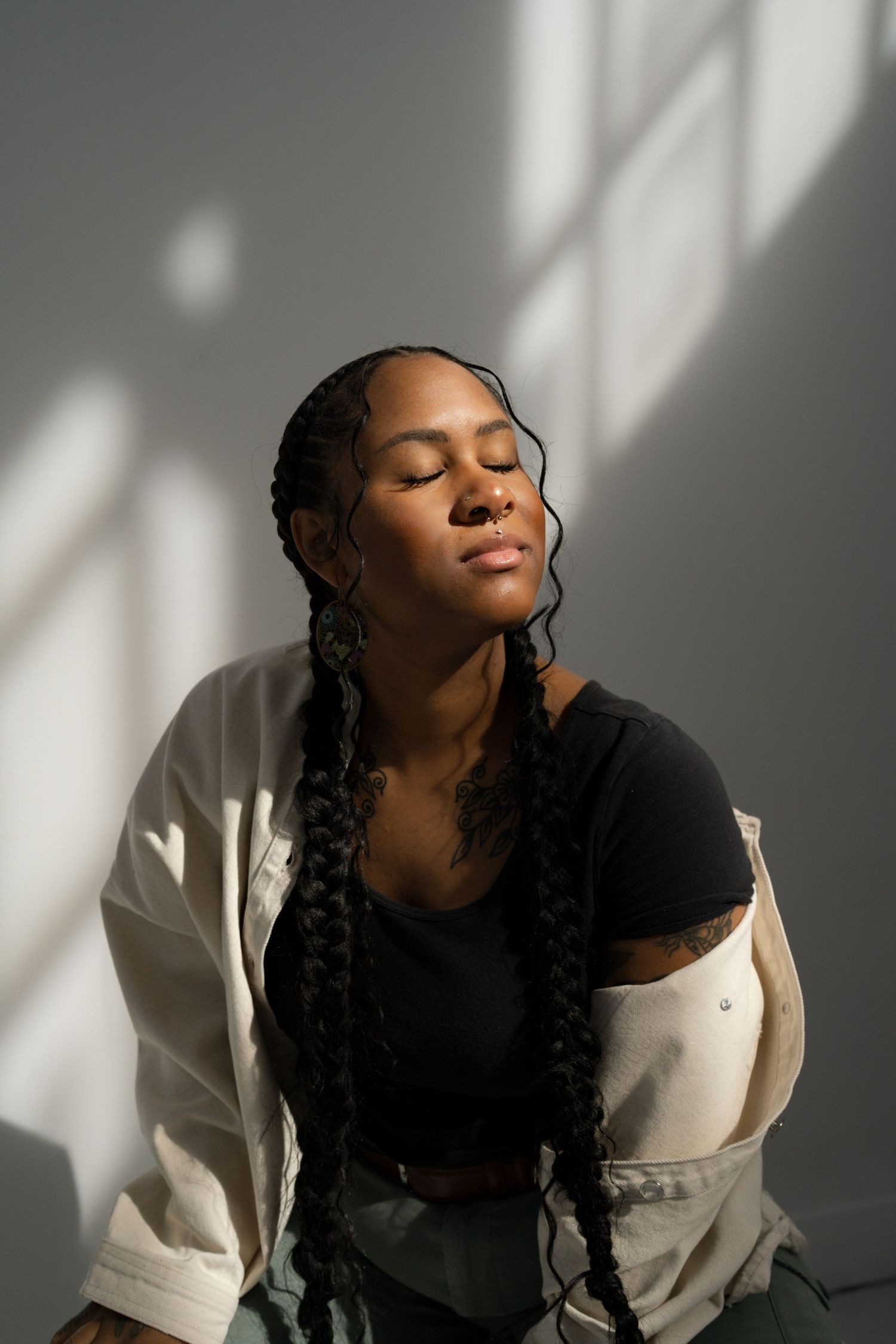 A Black woman with long braids closes her eyes peacefully in warm sunlight against a grey wall.