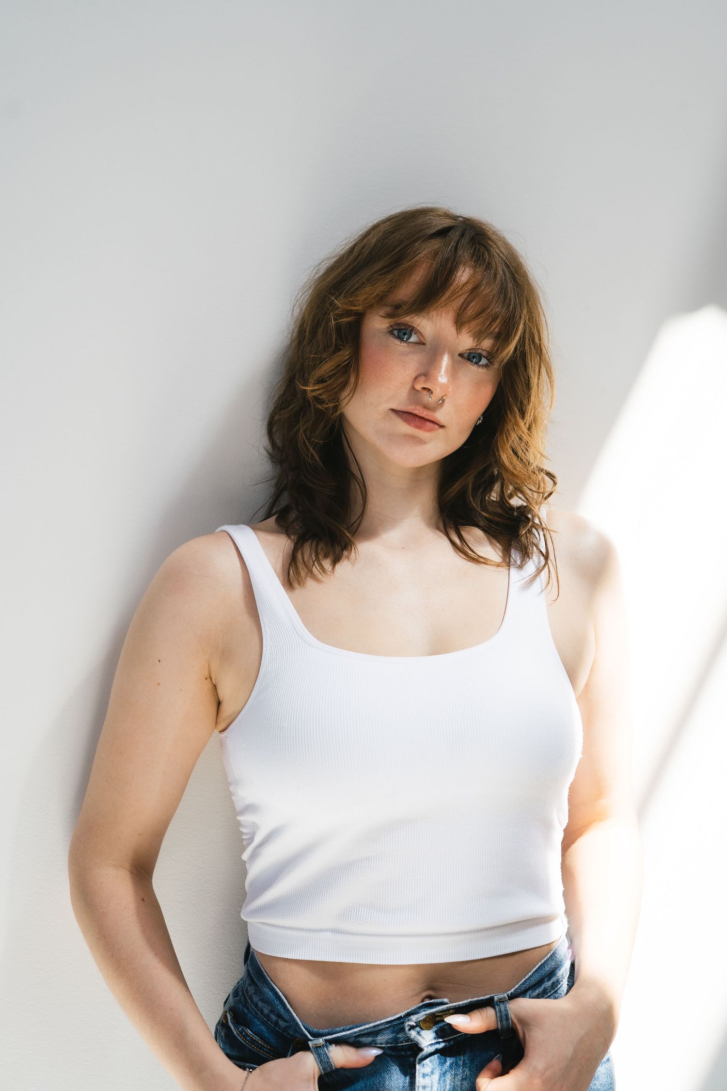 Young woman in white crop tank top and blue jeans leaning against white wall in natural light.