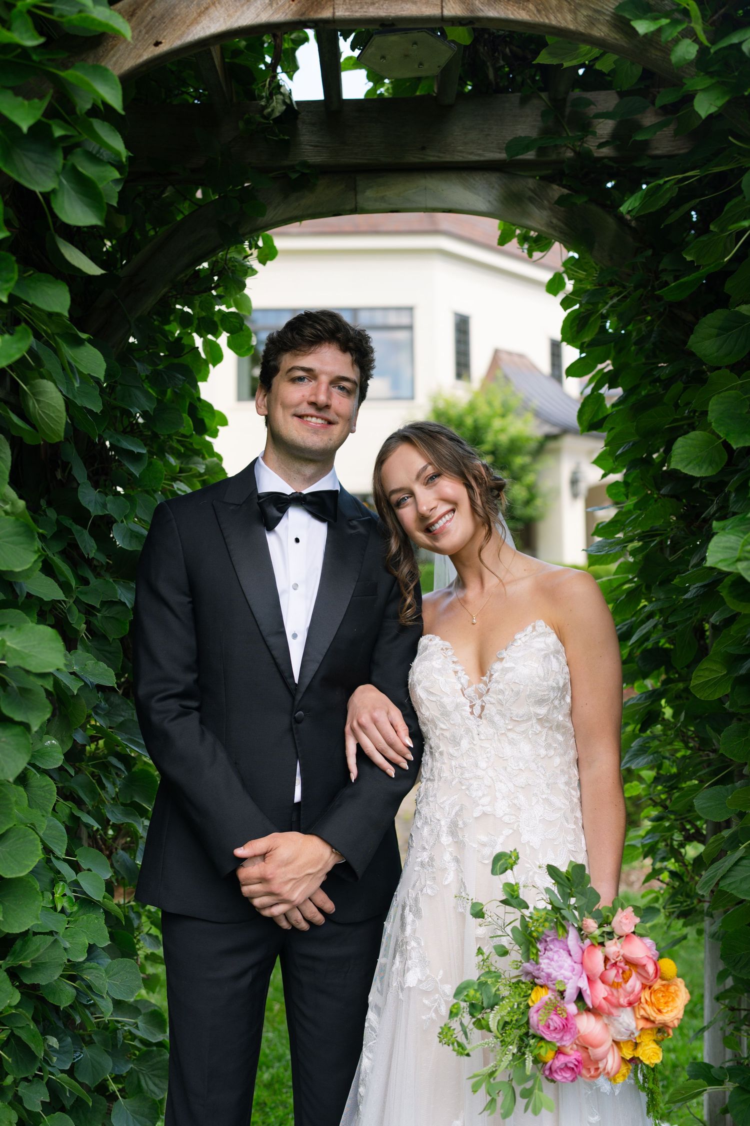 Bride and groom smile together under a leafy green archway, bride holding a colorful bouquet.