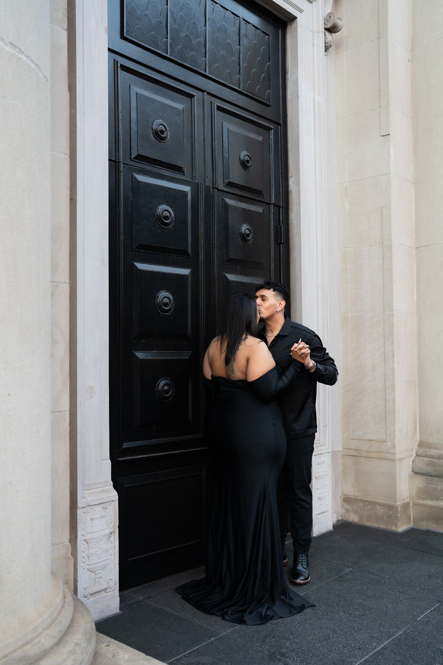 Couple sharing a kiss in matching black formal wear before an ornate black wooden door with stone columns.