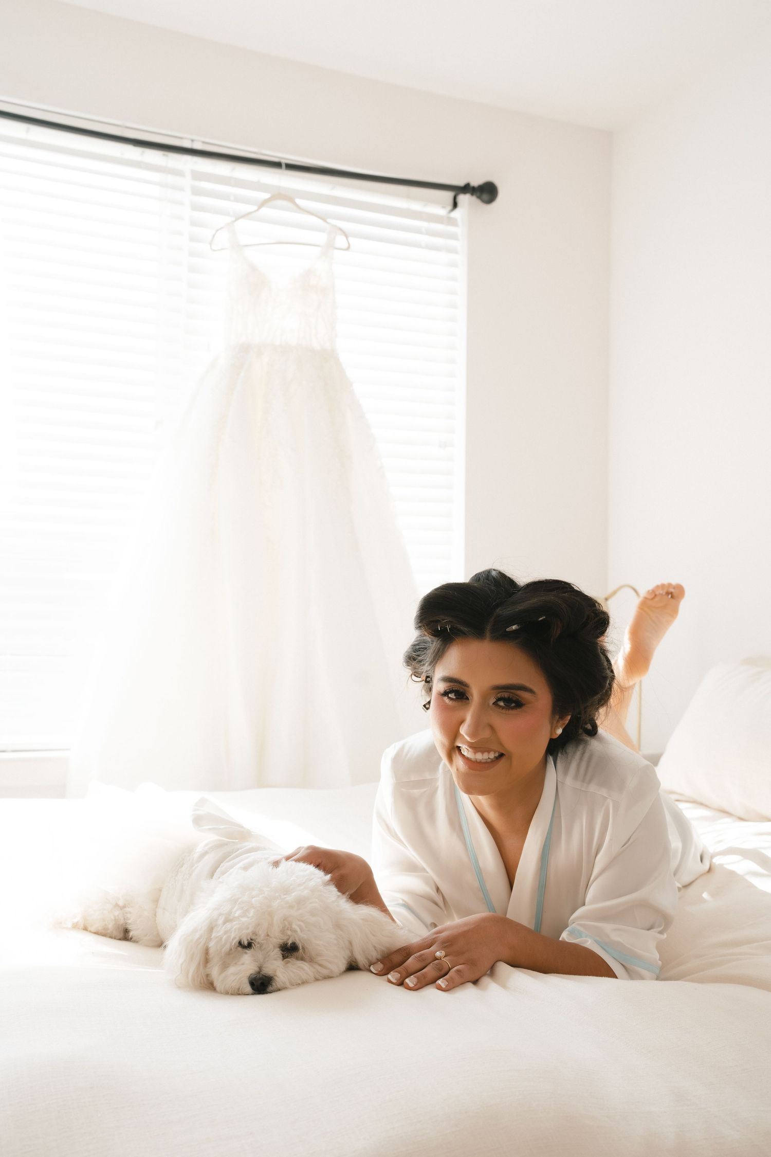 Smiling bride in white robe poses on bed with fluffy white dog, wedding gown hanging in background.