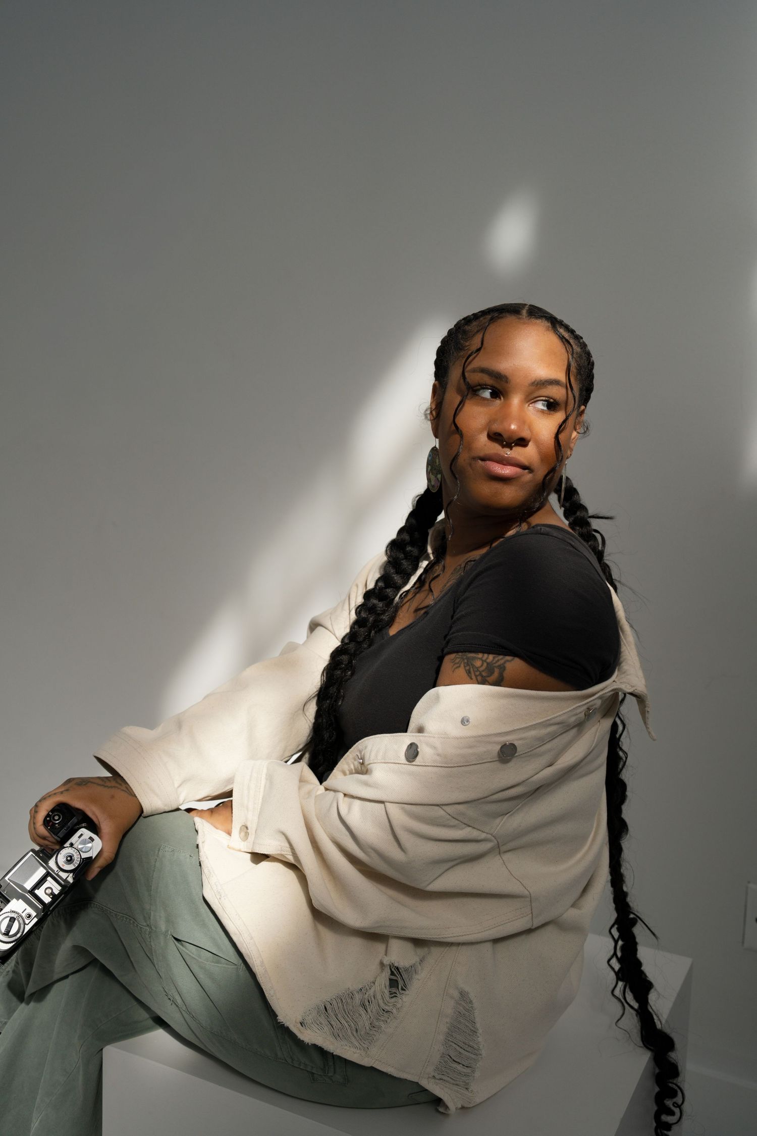 A Black woman with long braids sits on a white cube holding a camera, wearing a white jacket and sage green pants in a studio.