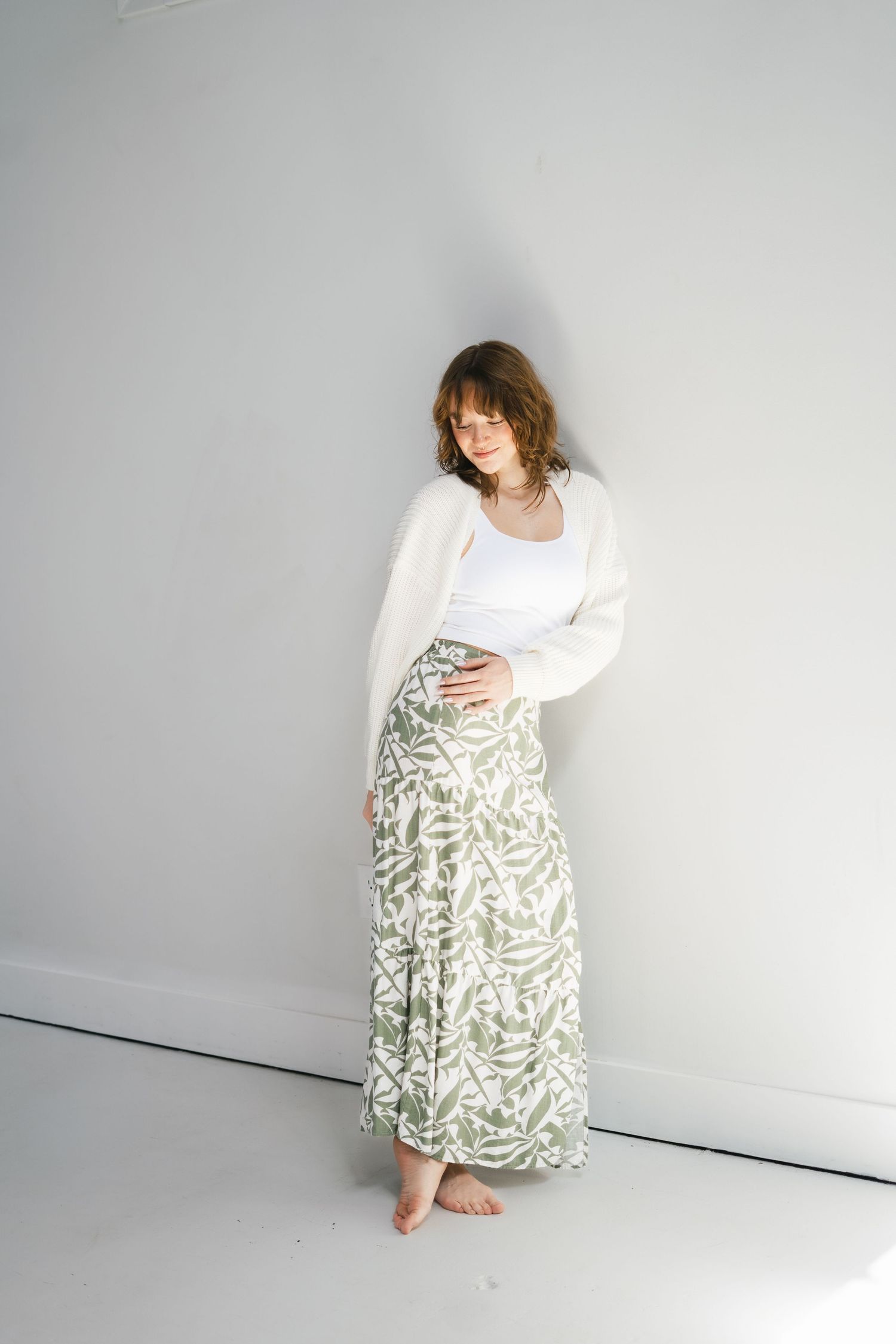 Pregnant woman in white top, cream cardigan, and sage green floral maxi skirt leaning against wall.