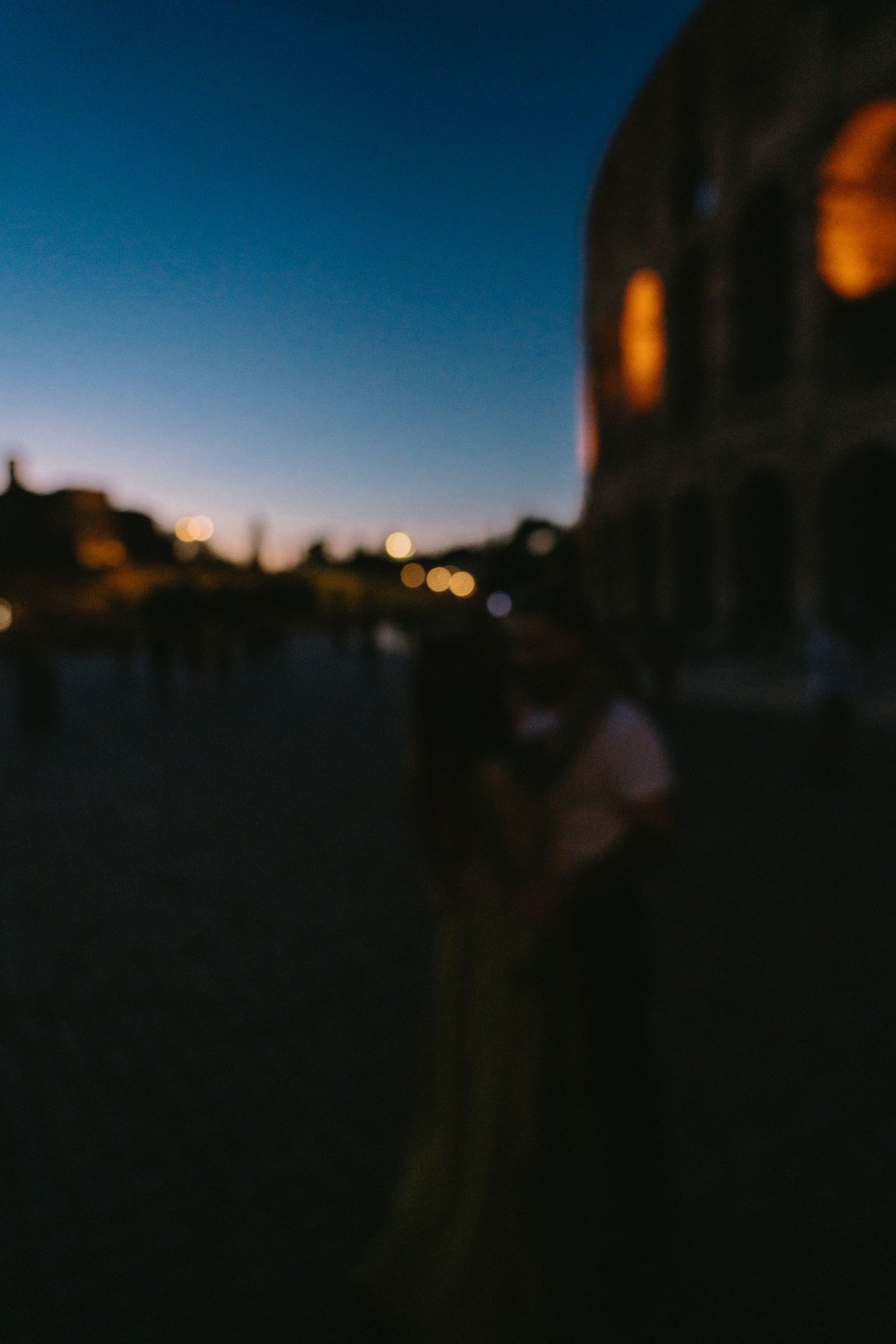 Blurry dusk scene near the Colosseum in Rome, with bokeh lights and silhouettes against a twilight sky.