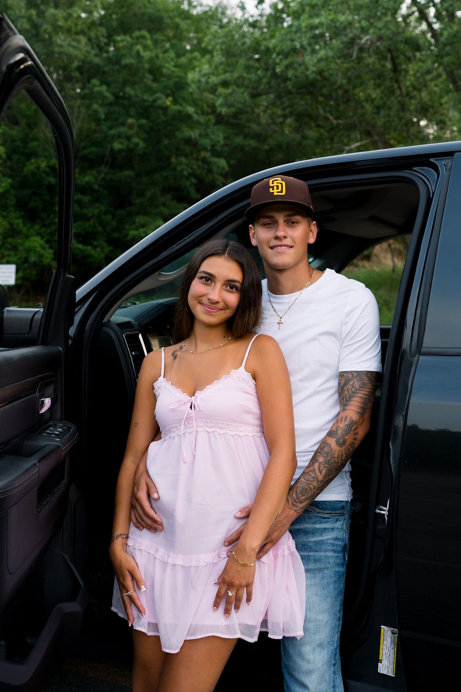Young couple posing by open black truck door, woman in pink dress, man in white tee with tattoos and SD cap.