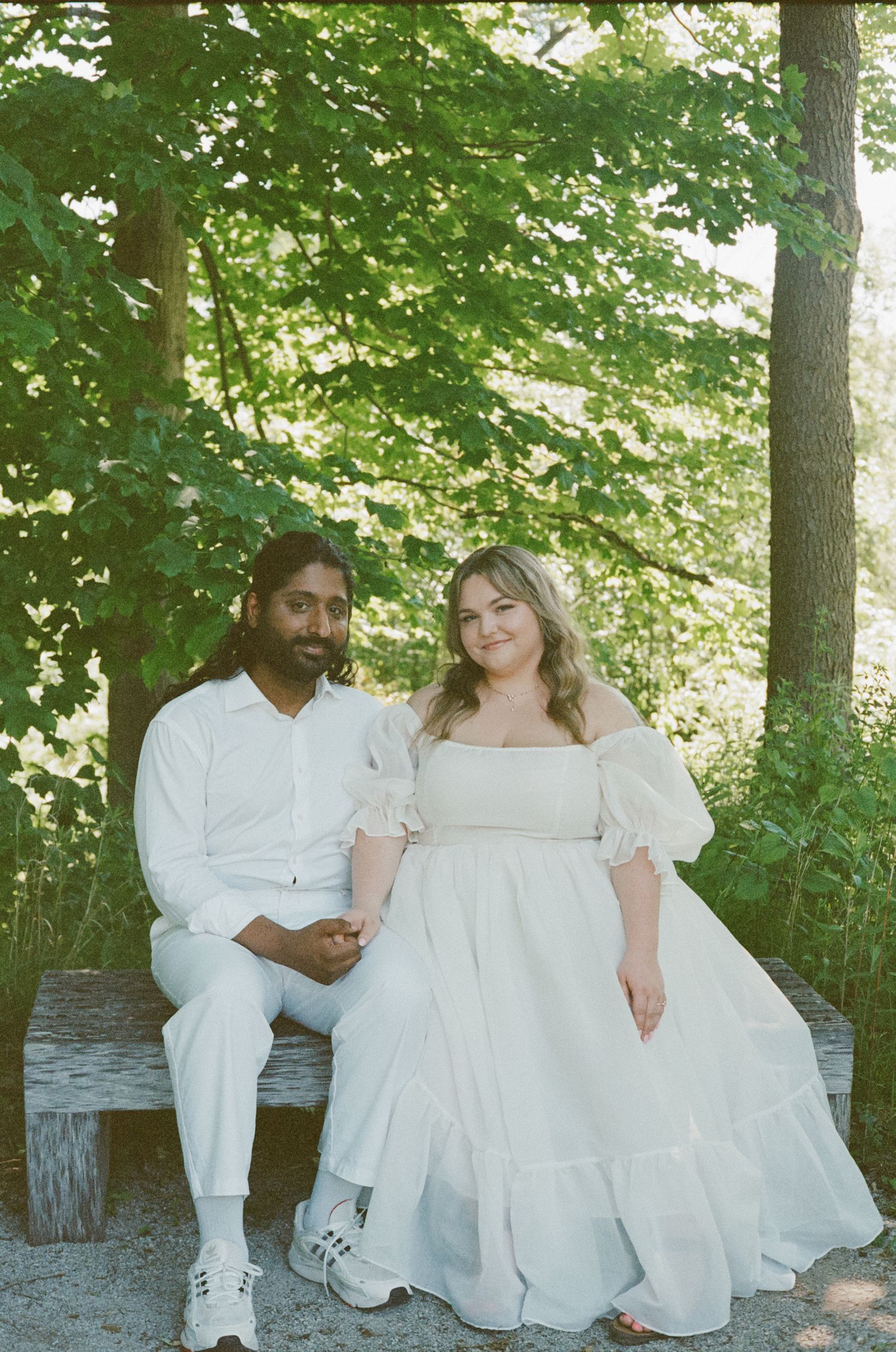 Couple sitting together on a bench in a lush green park, both dressed in white outfits.