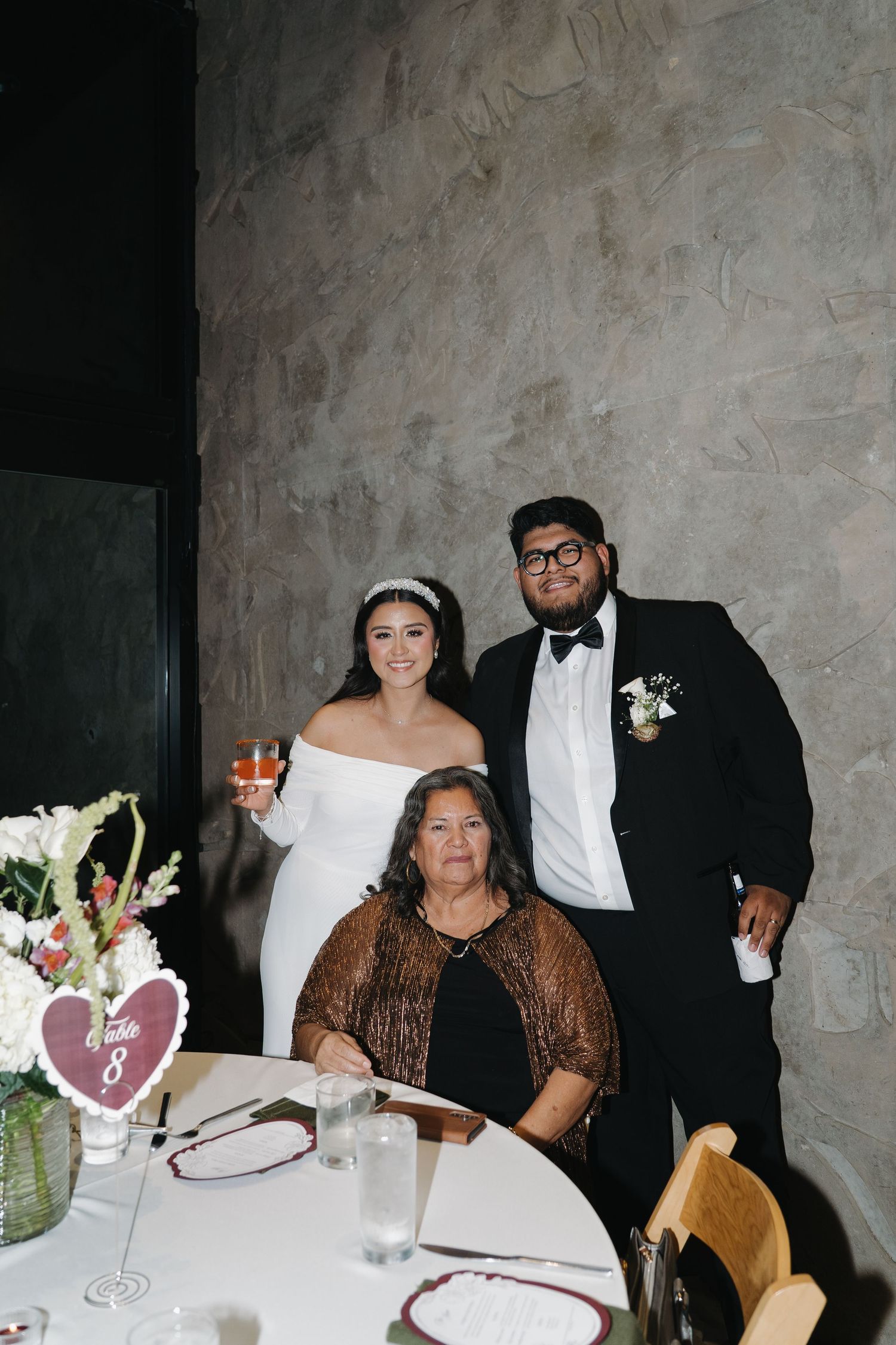 Bride and groom posing with seated elderly woman at wedding reception table number eight.