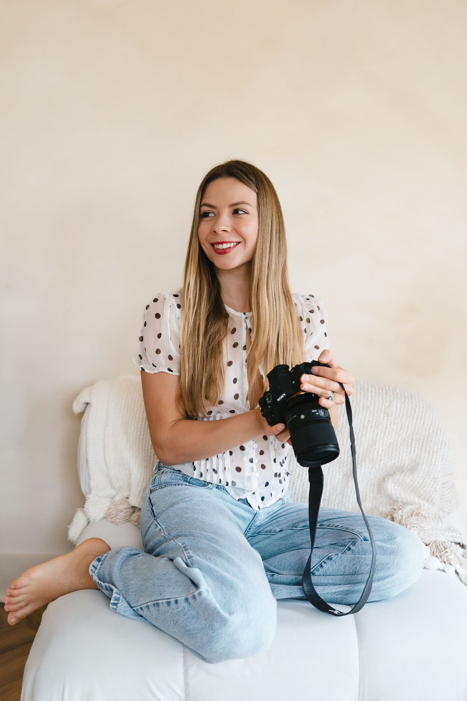 Smiling young woman in polka dot blouse and jeans sitting cross-legged on white sofa holding a camera.