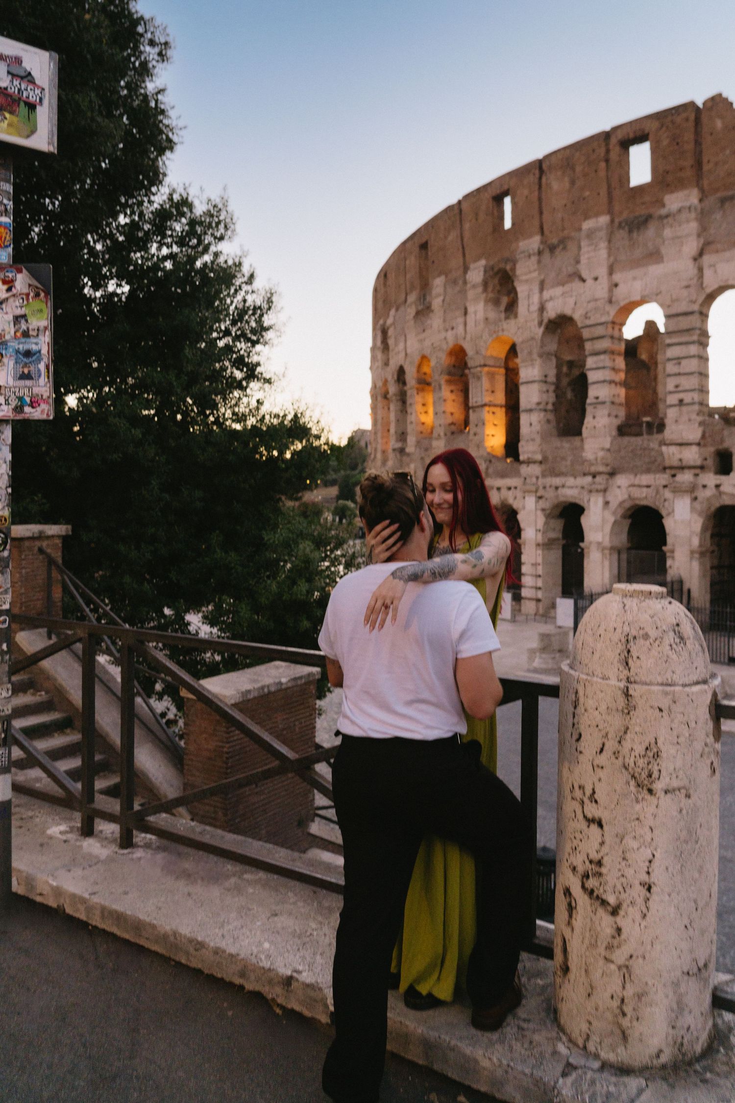 Couple embracing near the Colosseum in Rome at dusk, woman in green dress taking selfie.