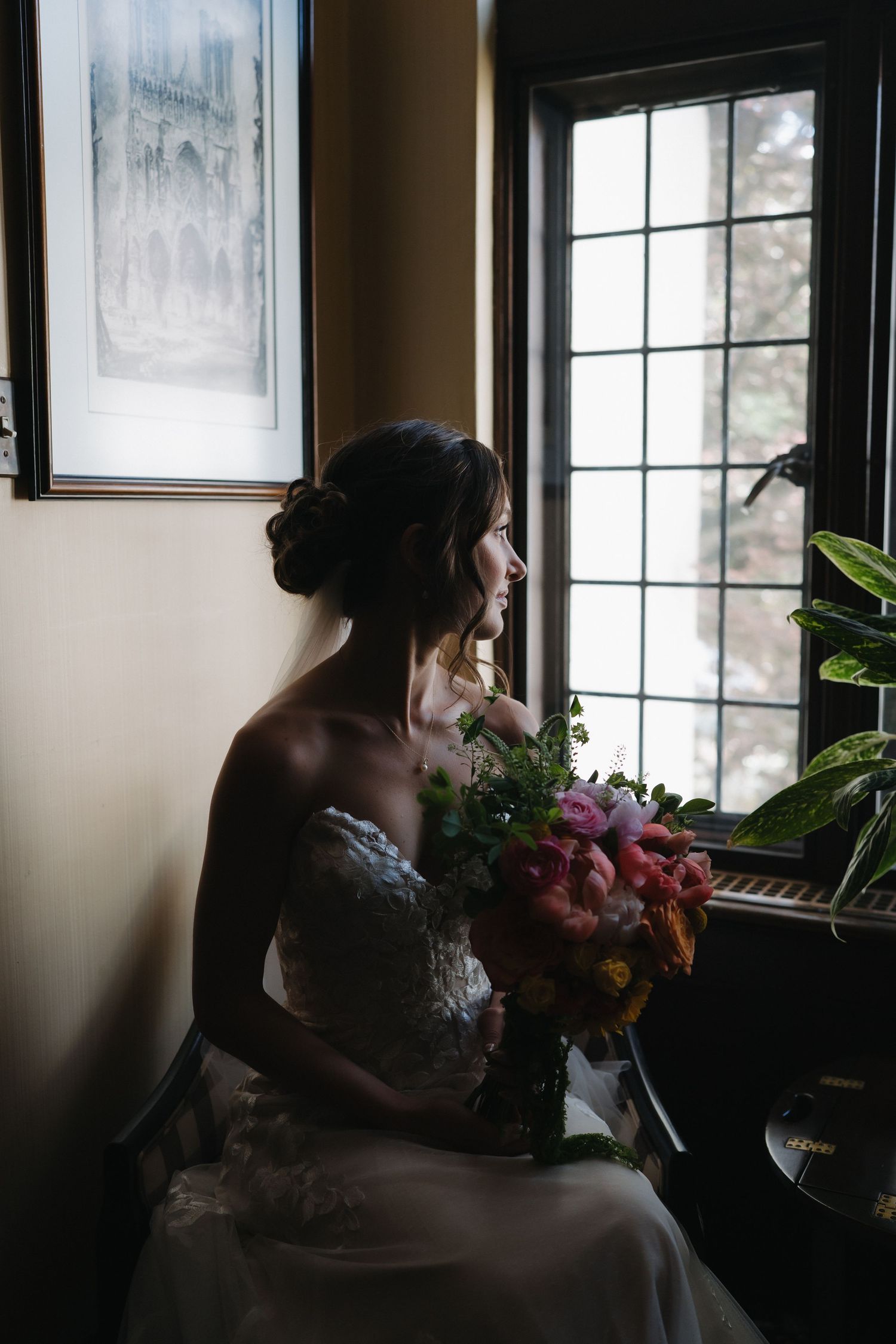 Bride in lace wedding gown sits by a window holding a colorful bouquet in soft natural light.