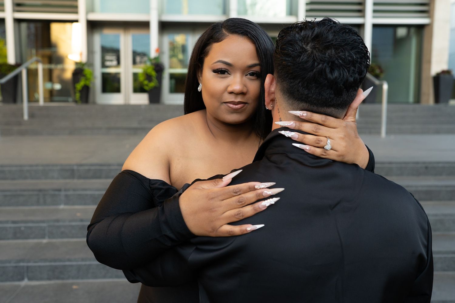Woman in black off-shoulder dress embracing a man on steps outside a modern building.