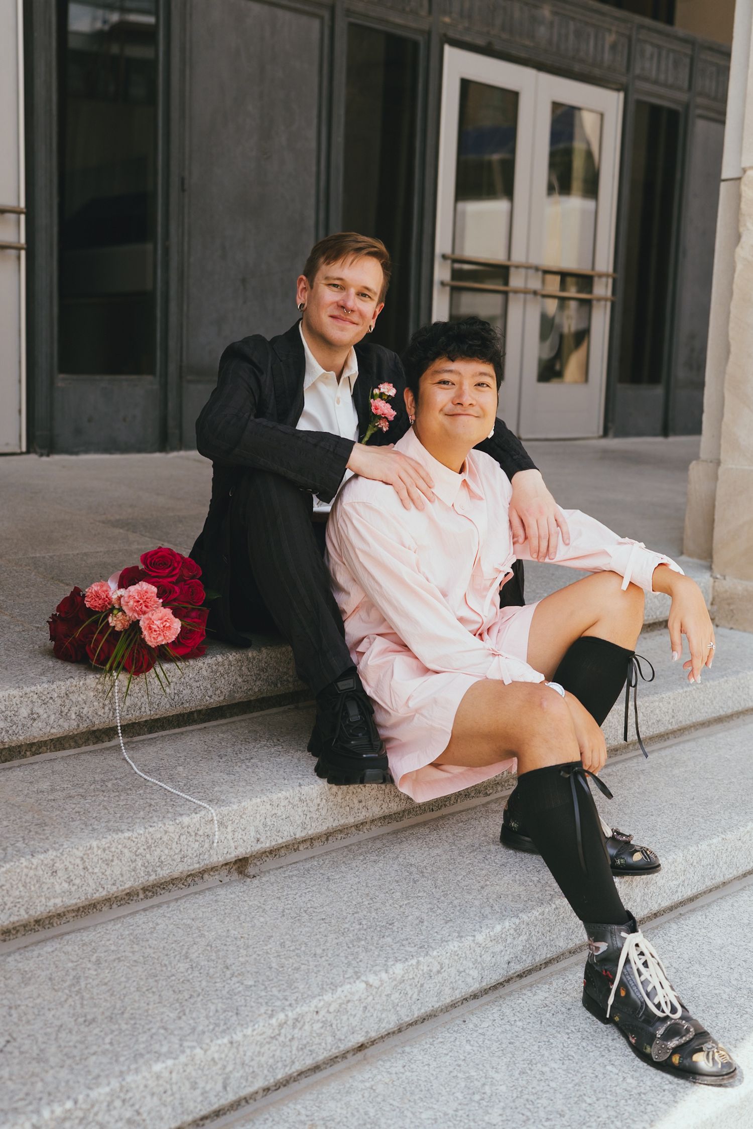 Couple sitting on stone steps smiling, man in dark suit with boutonniere, woman in pink dress with black boots.