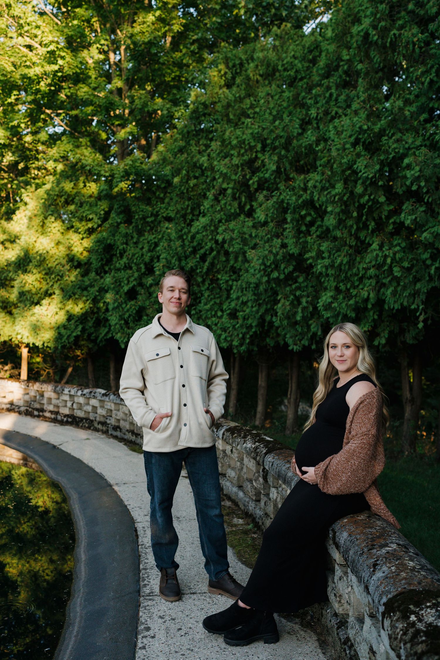 Pregnant couple posing by a curved stone wall path with tall green trees in soft evening light.