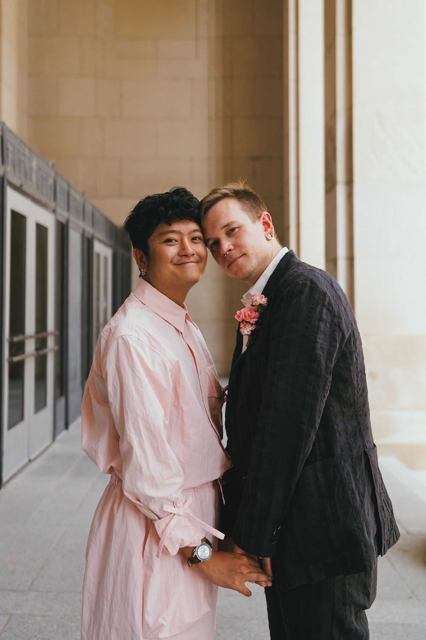 Interracial couple posing together, woman in pink dress and man in dark suit with floral boutonniere, smiling warmly.
