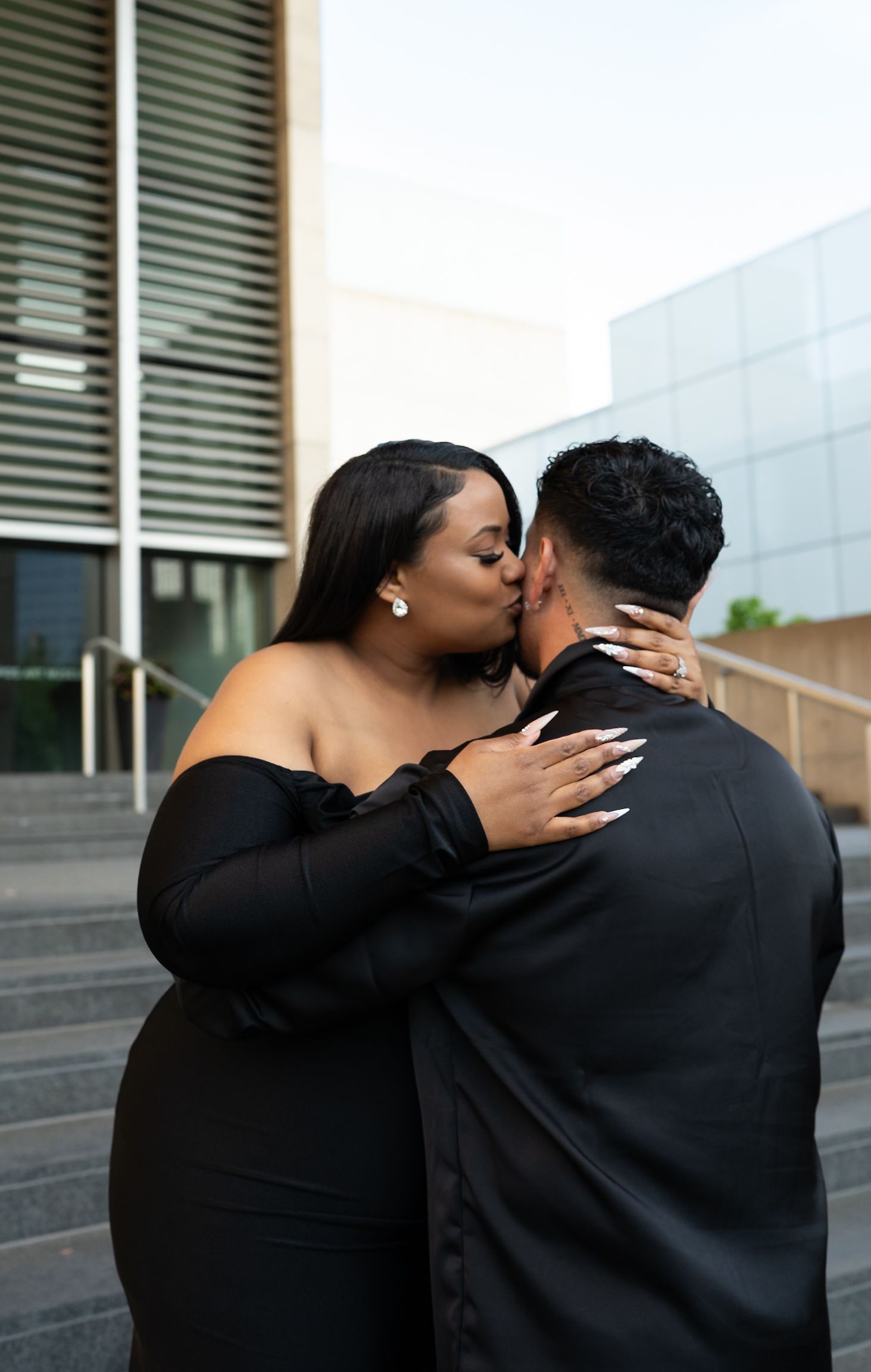 Couple sharing a romantic kiss outdoors on steps while dressed in elegant black formal attire.