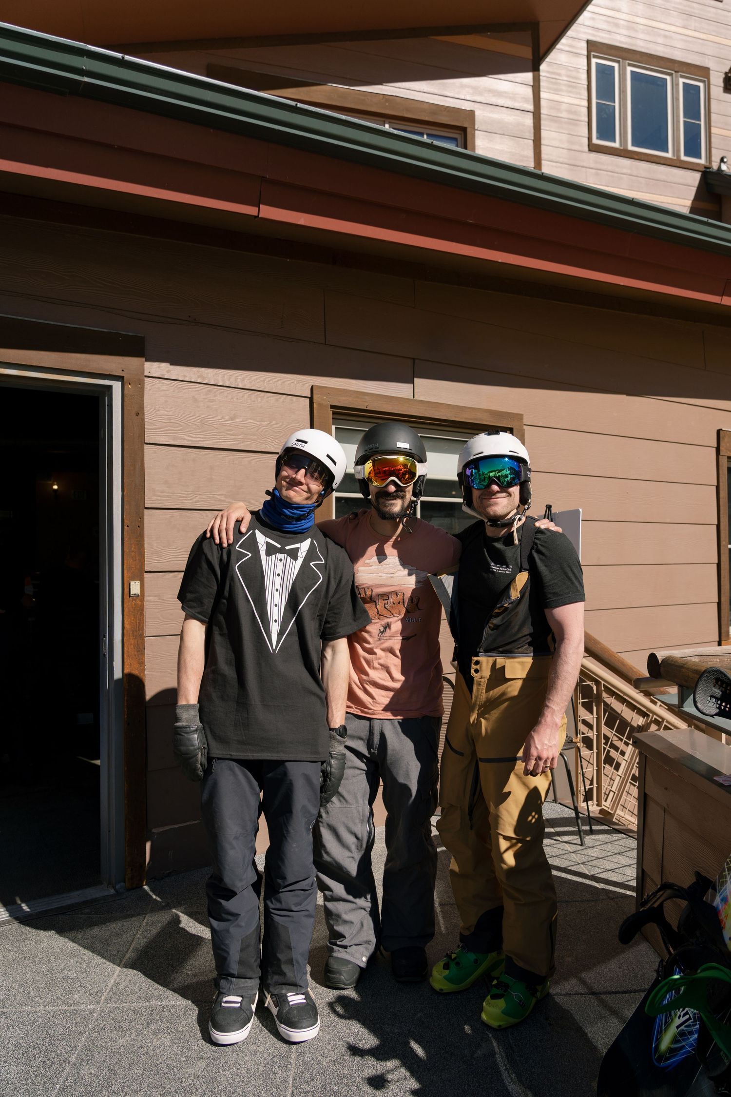 Three people wearing helmets and ski gear posing together outside a mountain lodge building.