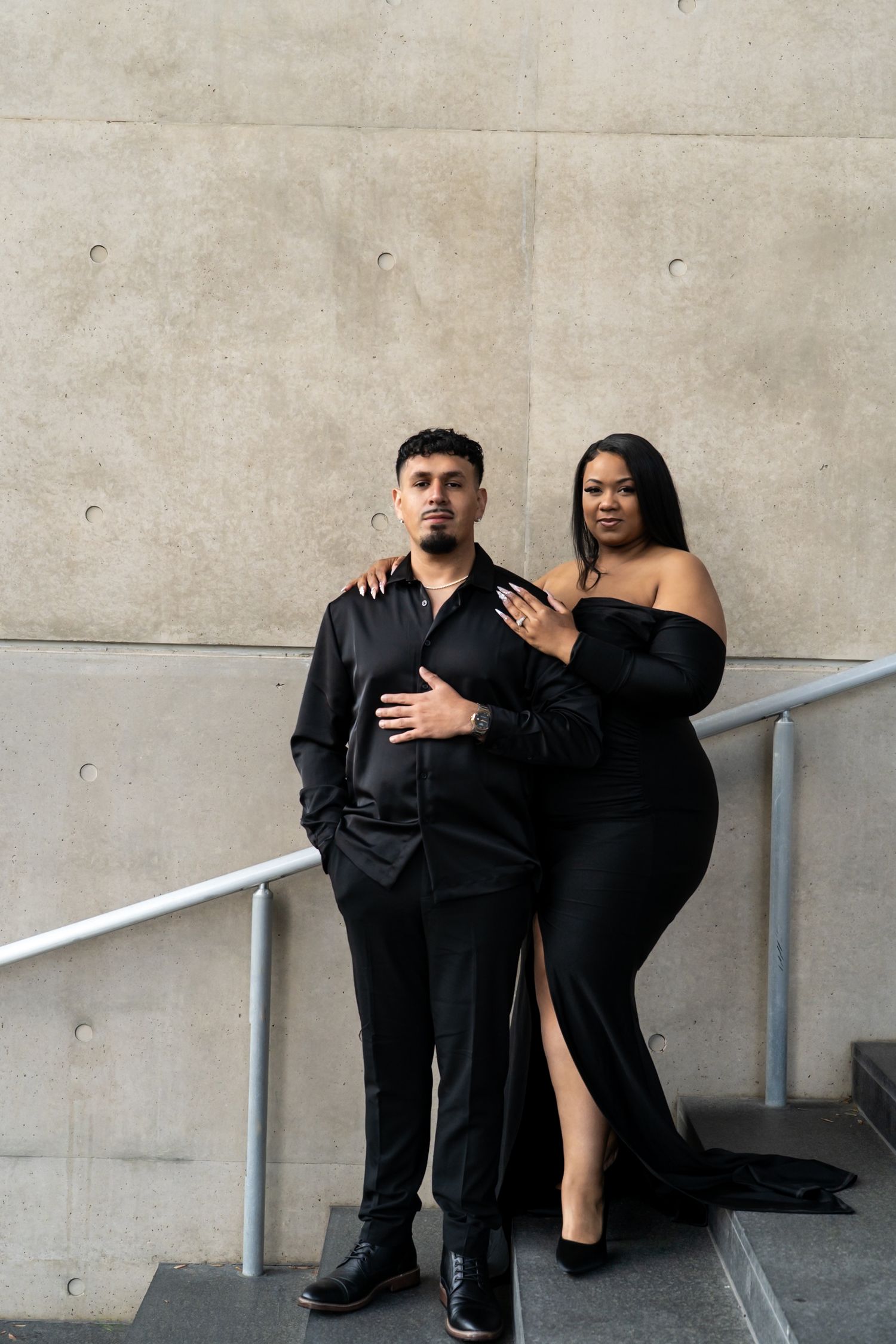 Couple in matching black formal wear posing on steps against concrete wall, woman in off-shoulder gown, man in black shirt.