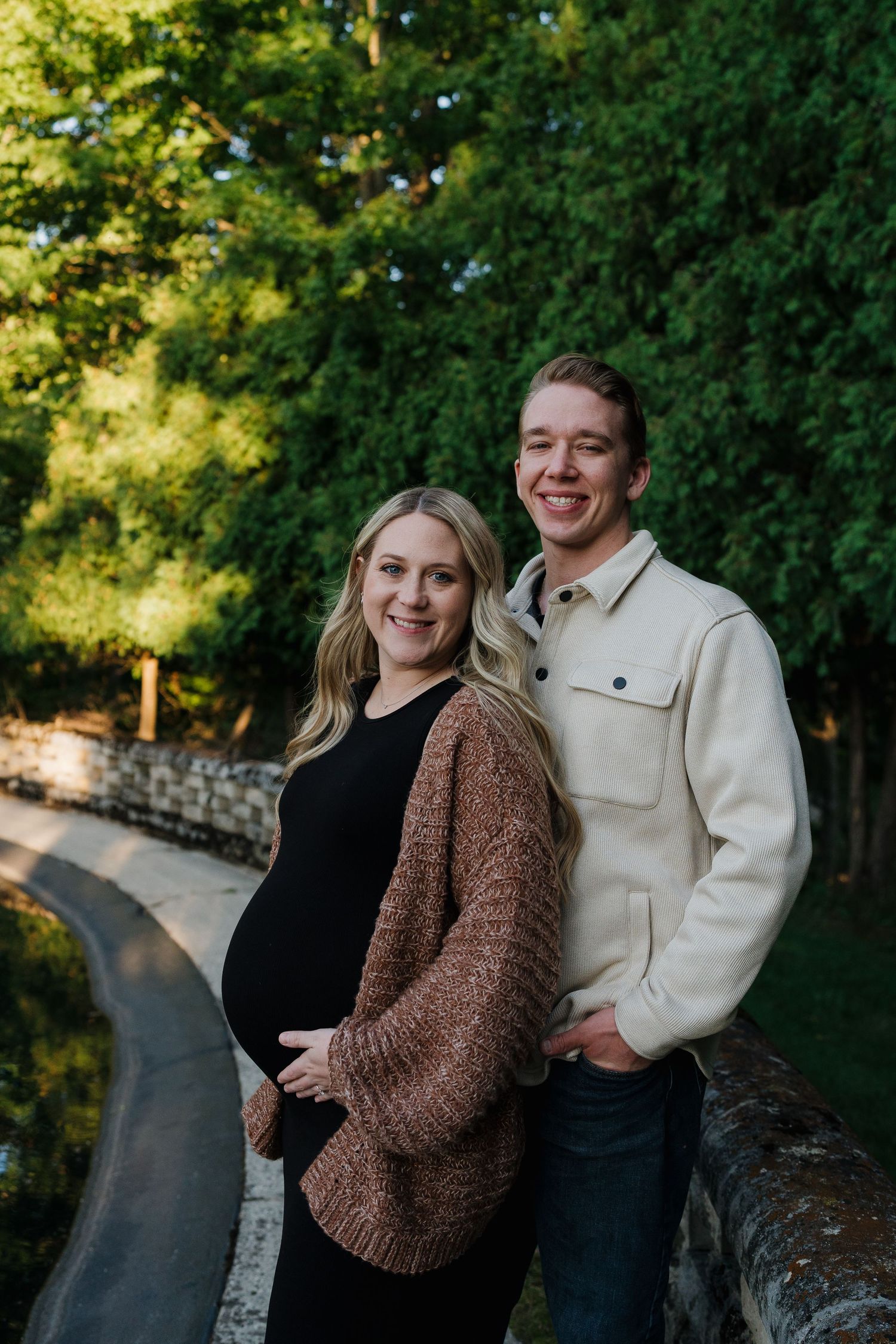 Smiling pregnant couple posing on a stone bridge surrounded by lush green trees.