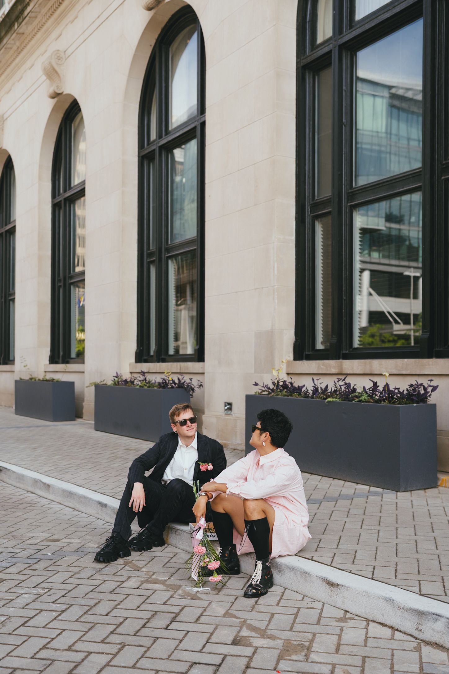 Newlywed couple in sunglasses relaxing on steps outside grand stone building with flowers.