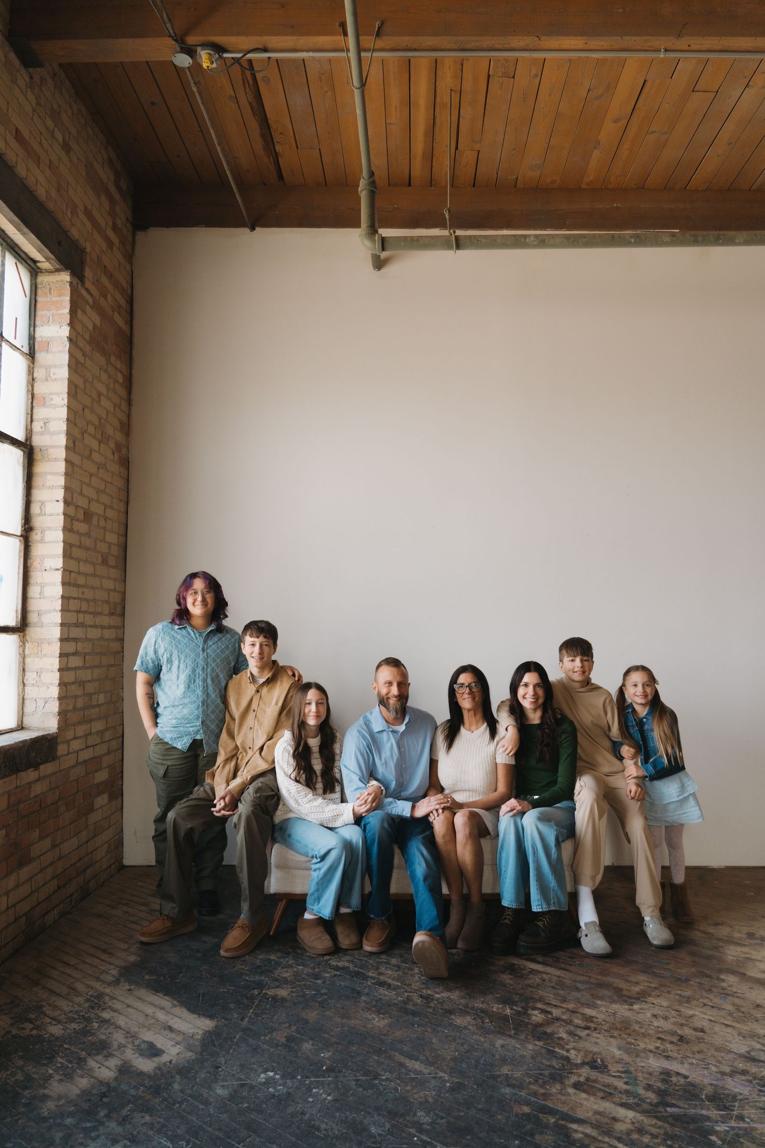Large family group portrait with seven people posing together in a rustic brick studio with wood ceiling.