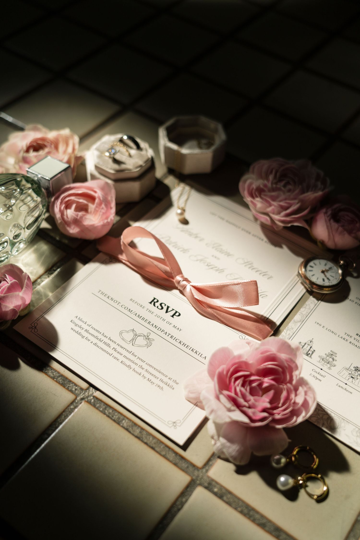 Elegant wedding RSVP invitation surrounded by pink roses, jewelry, and vintage clock on tile surface.