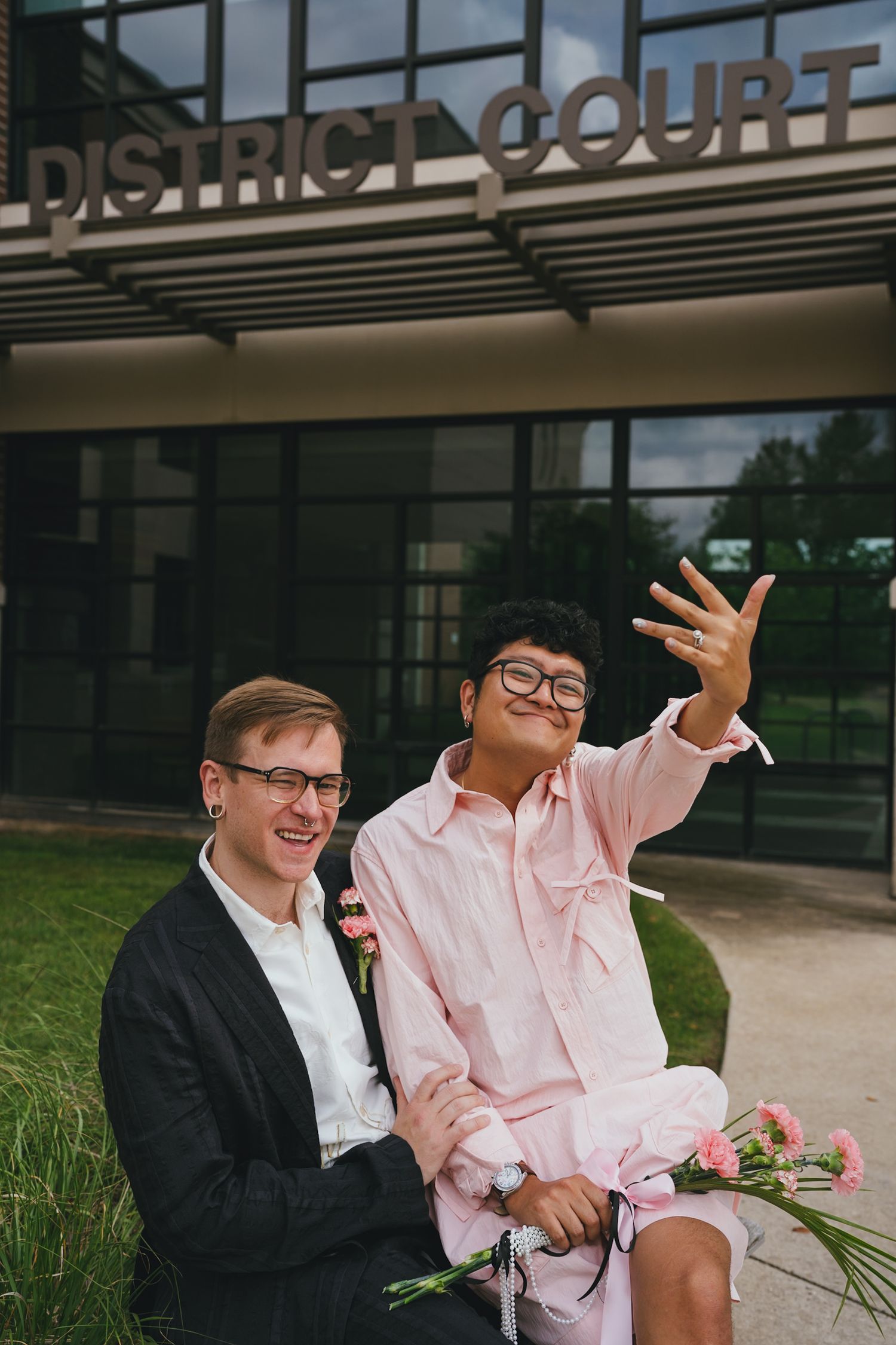 Happy newlywed couple showing off wedding ring outside District Court building with pink flowers.