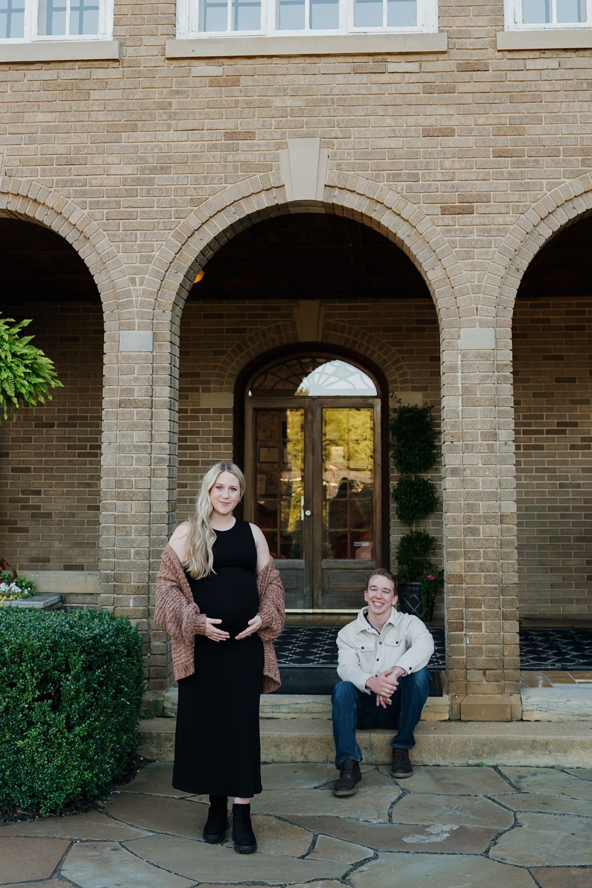 A pregnant woman in black dress poses with partner outside a brick building with arched columns.