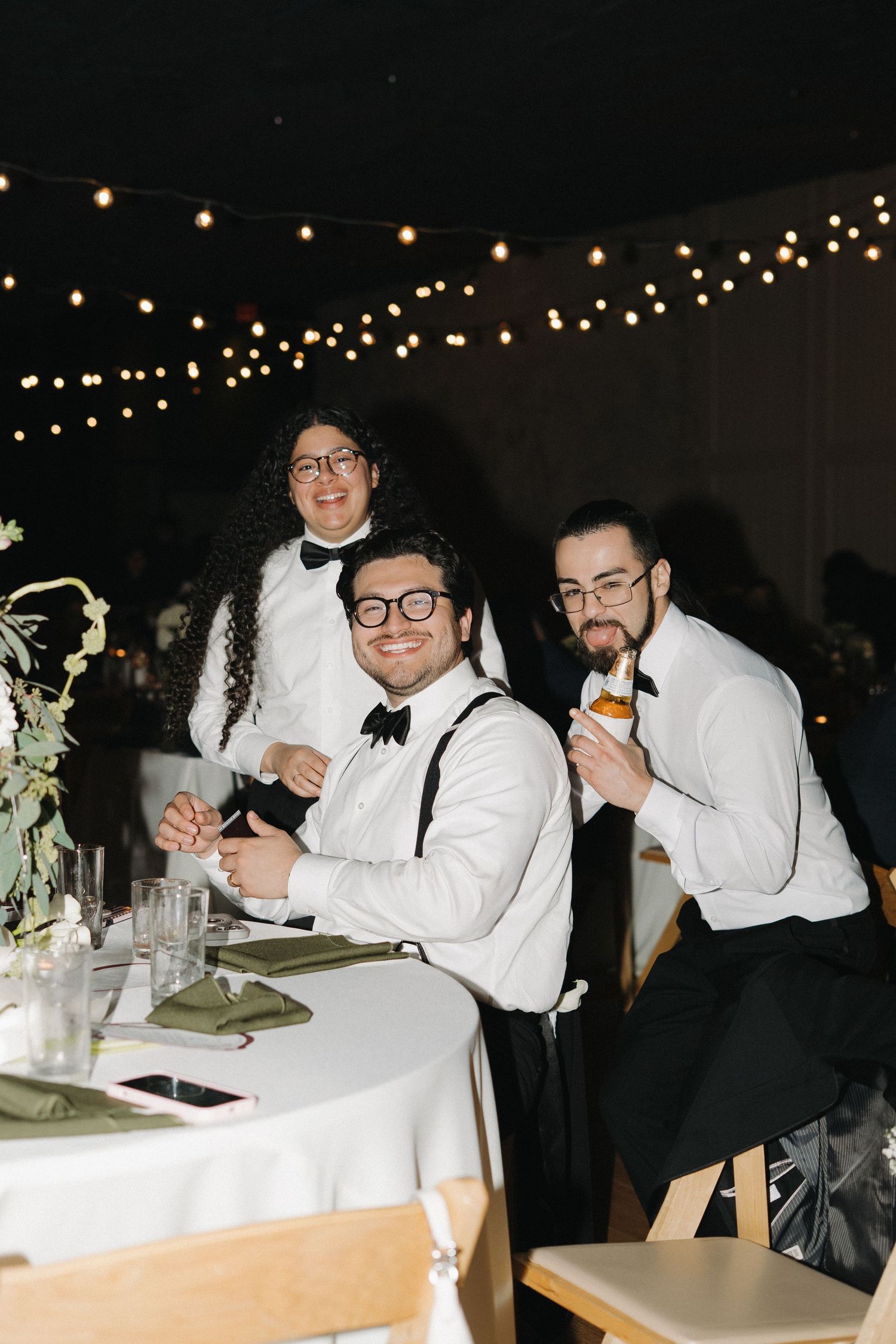 Three groomsmen in white shirts and bow ties smiling at wedding reception table with string lights.
