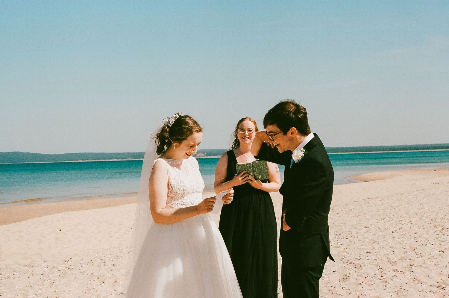 Bride and groom exchanging rings on a beach with officiant under blue sky.