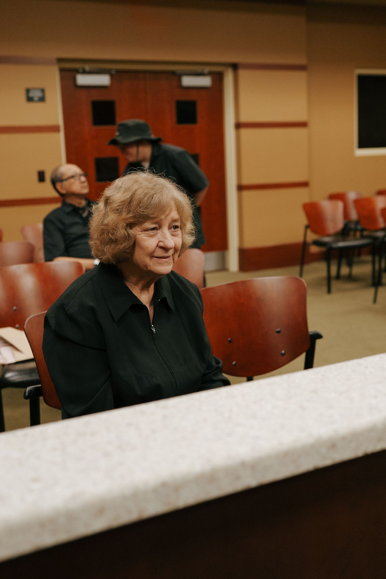 Elderly woman in black sitting in red chairs inside a courthouse waiting room.