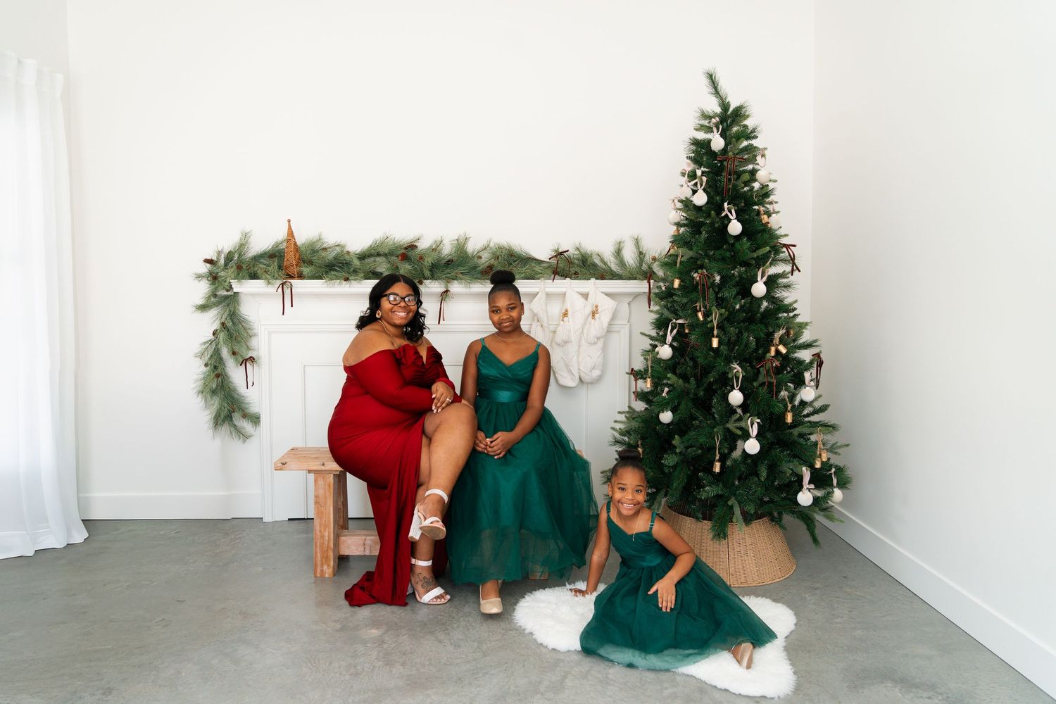 Mother and two daughters in red and green holiday dresses pose playfully near a Christmas tree.